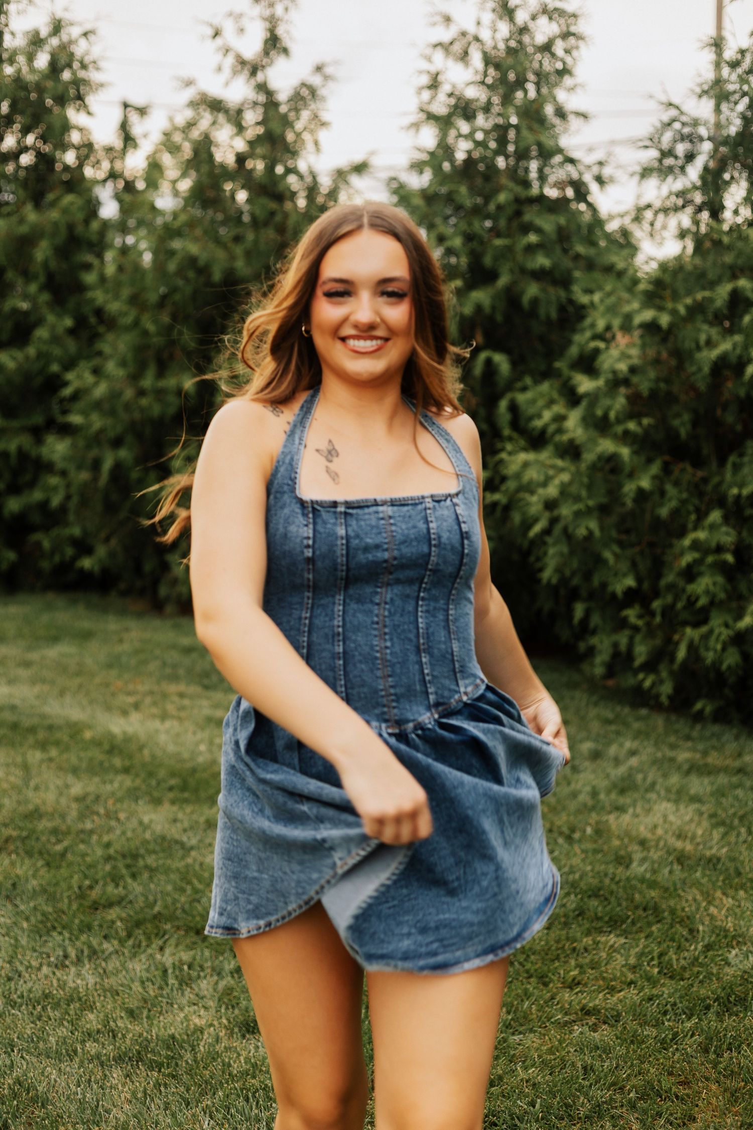 Woman in denim dress poses playfully on grass with evergreen trees in background.