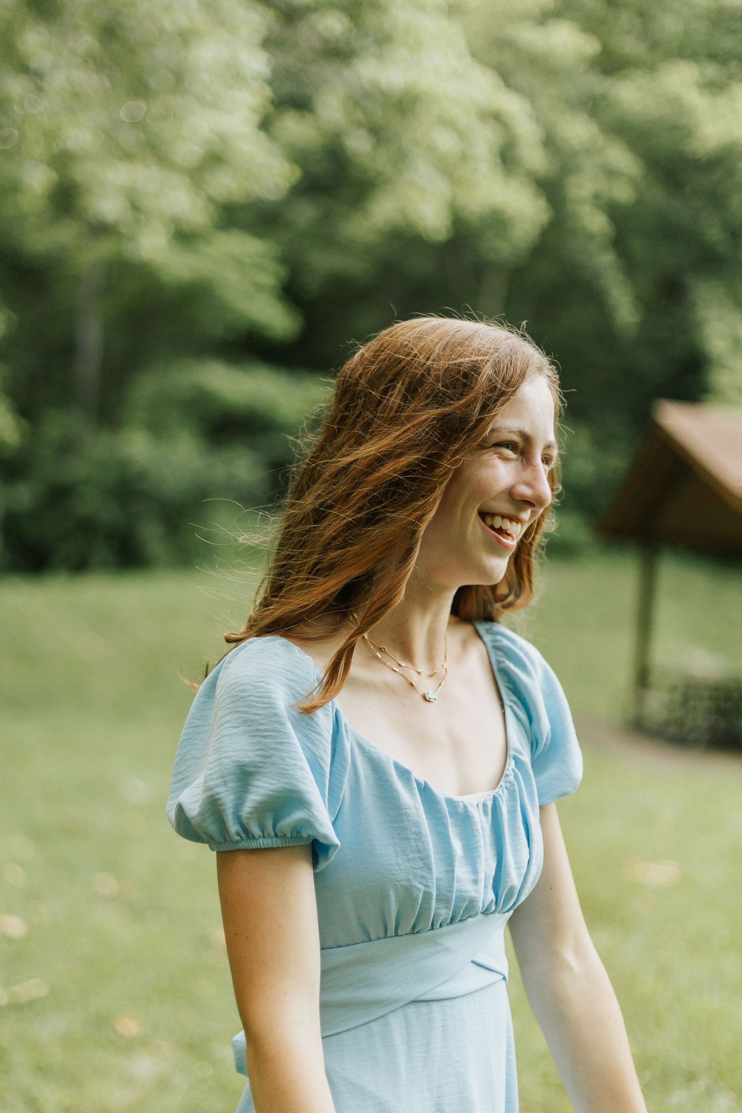 Young woman in light blue dress smiling outdoors in a sunny garden setting.