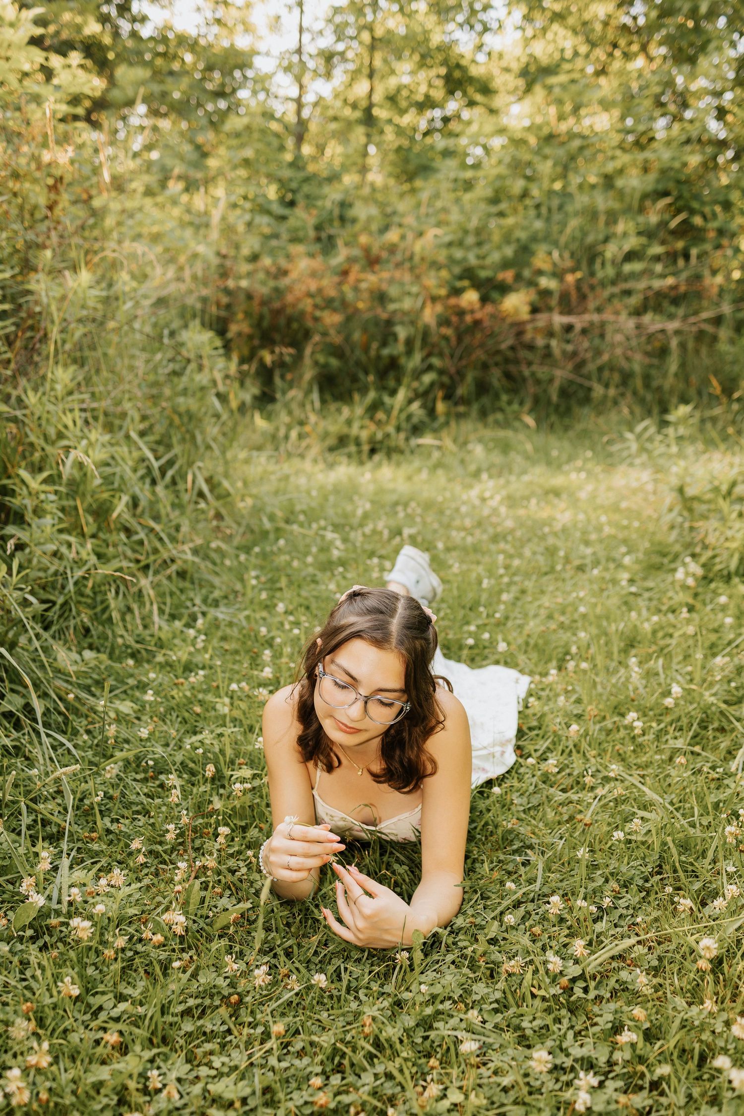 Young woman with glasses lying on green grass in a meadow, holding a wildflower, surrounded by lush trees.
