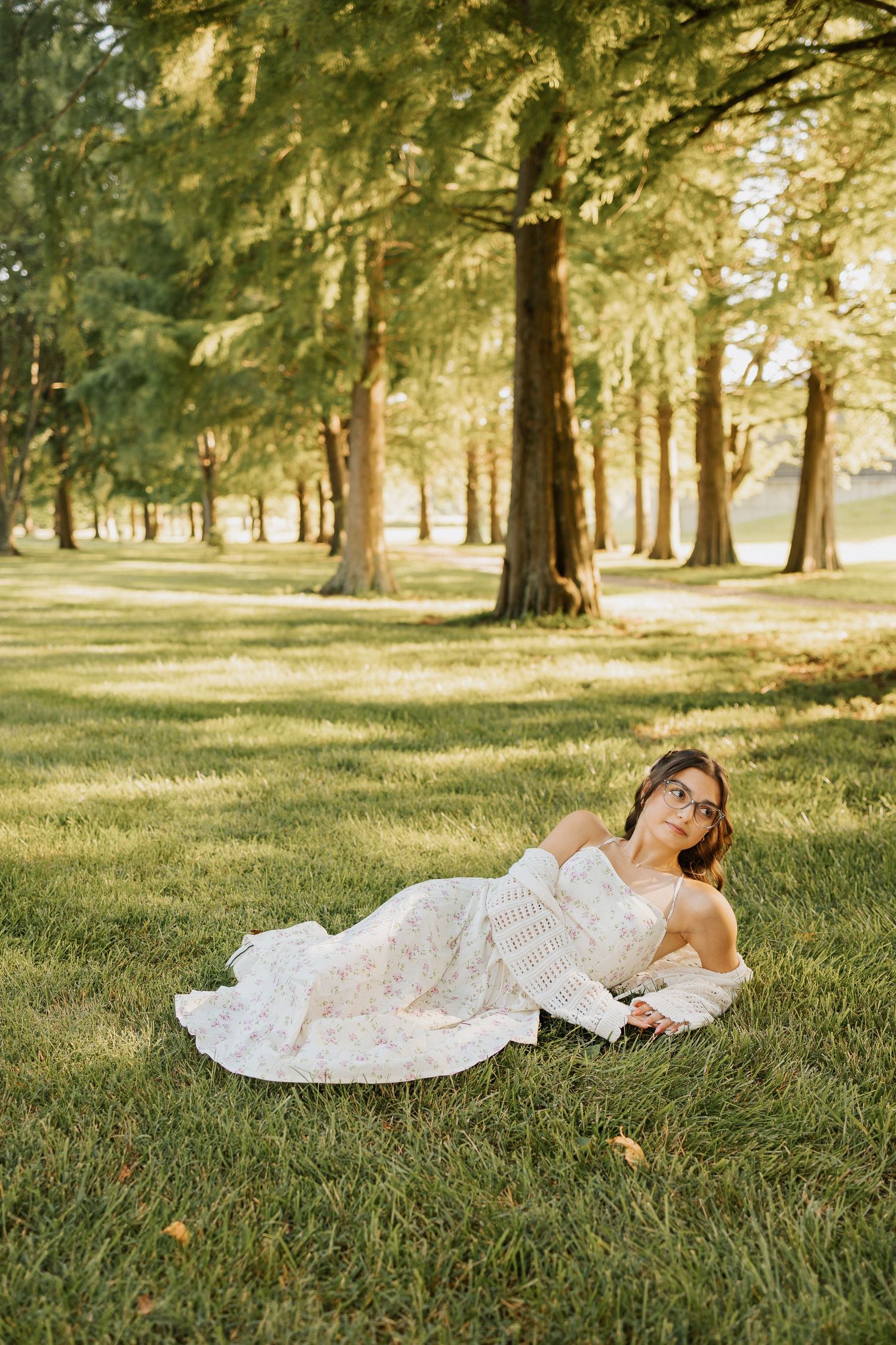 A bride in a floral gown reclines on lush green grass surrounded by tall trees in a sunlit park.