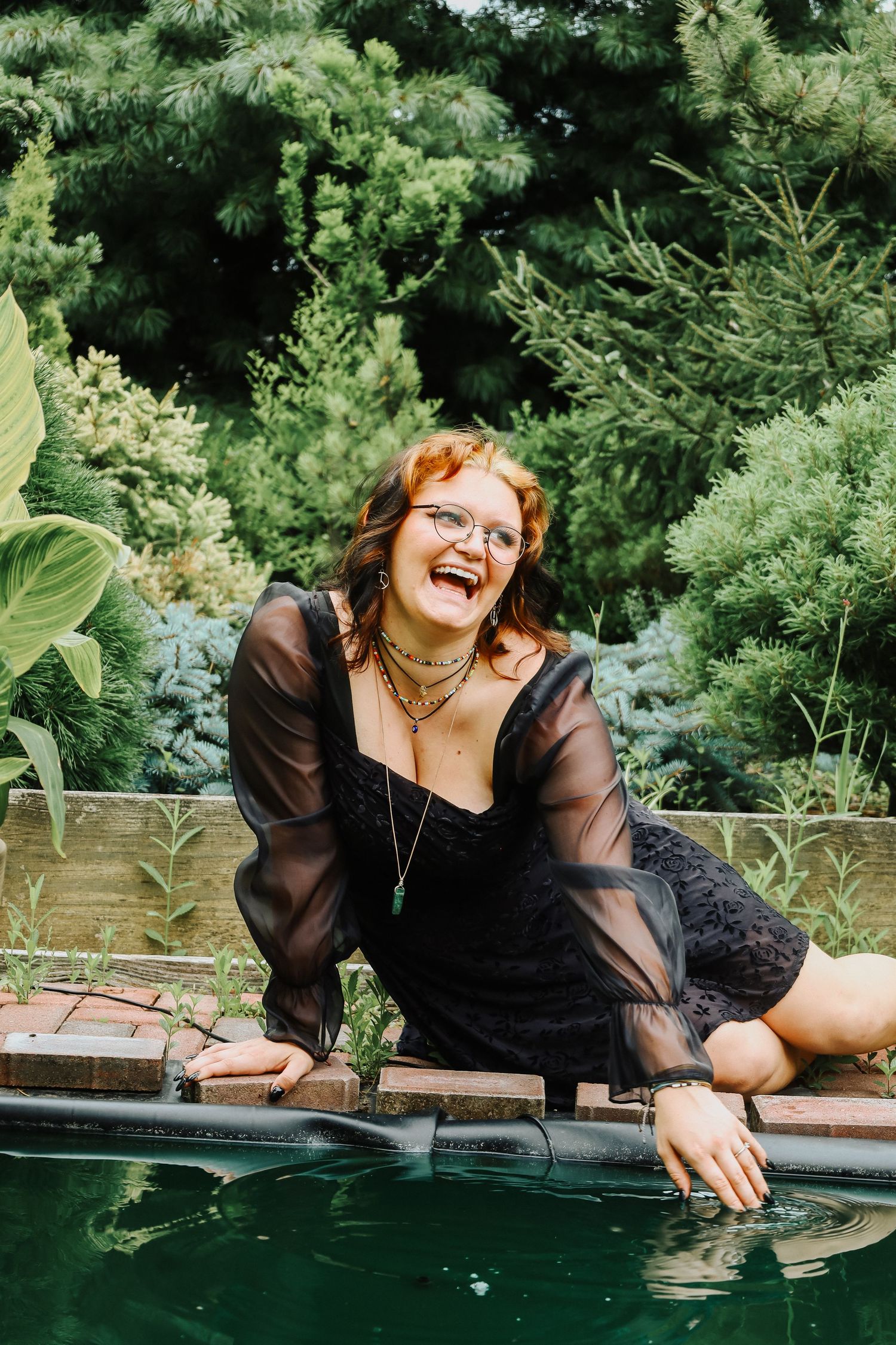 Person in black dress laughing while leaning on reflective surface near garden greenery.