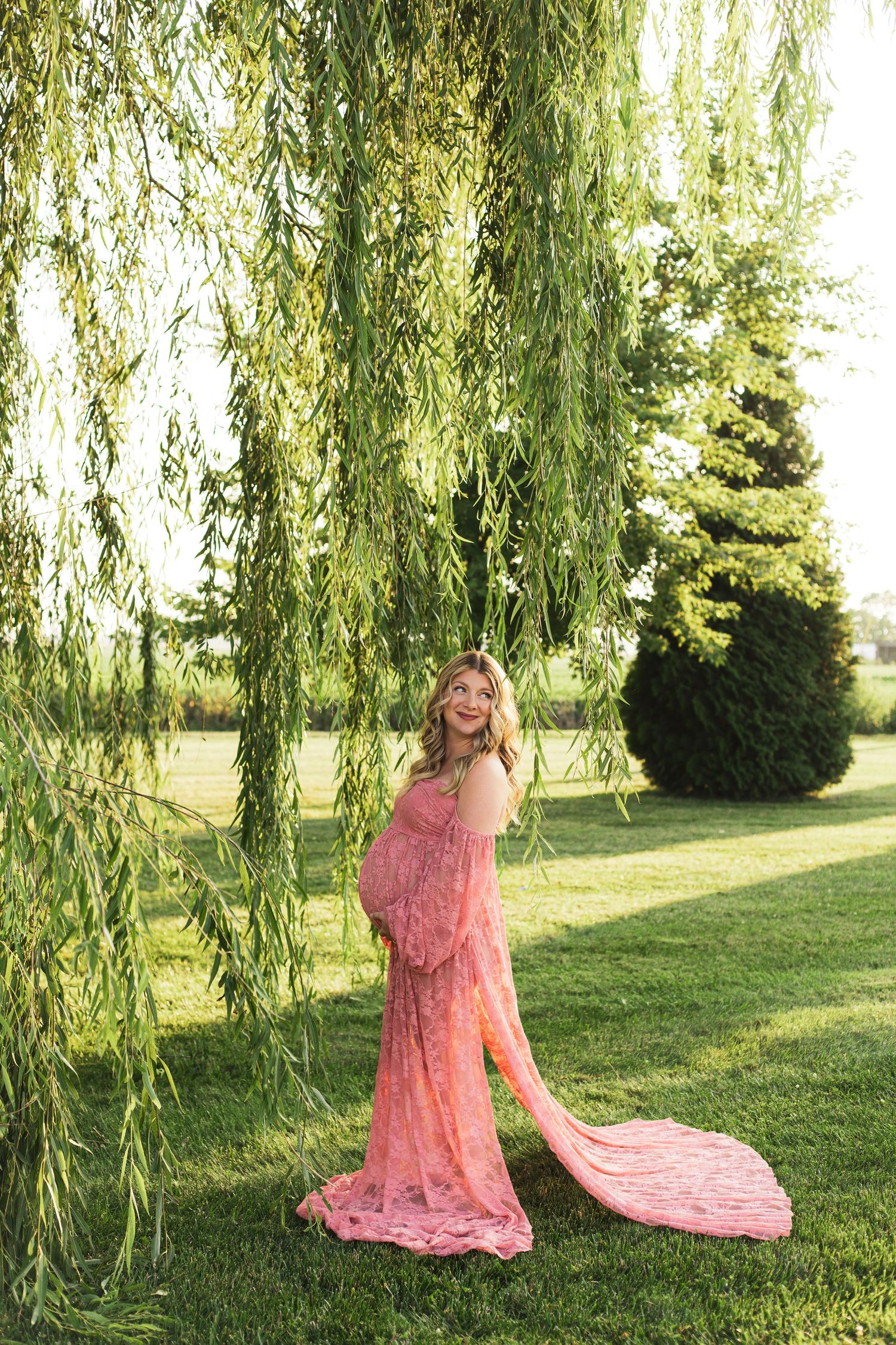 A maternity photoshoot under a weeping willow tree featuring a flowing coral lace dress.