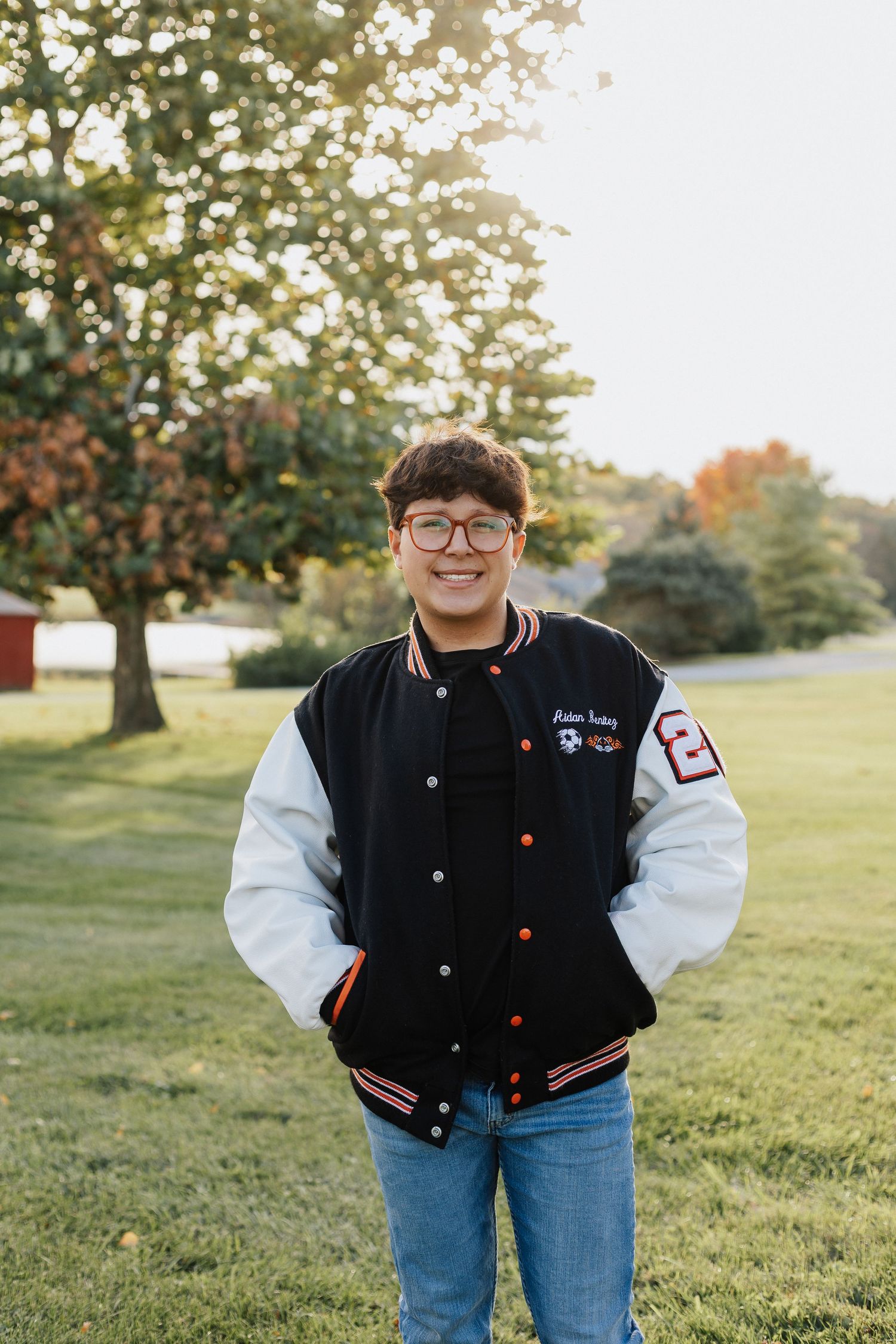 Person wearing varsity letterman jacket and jeans poses in grassy field at sunset.