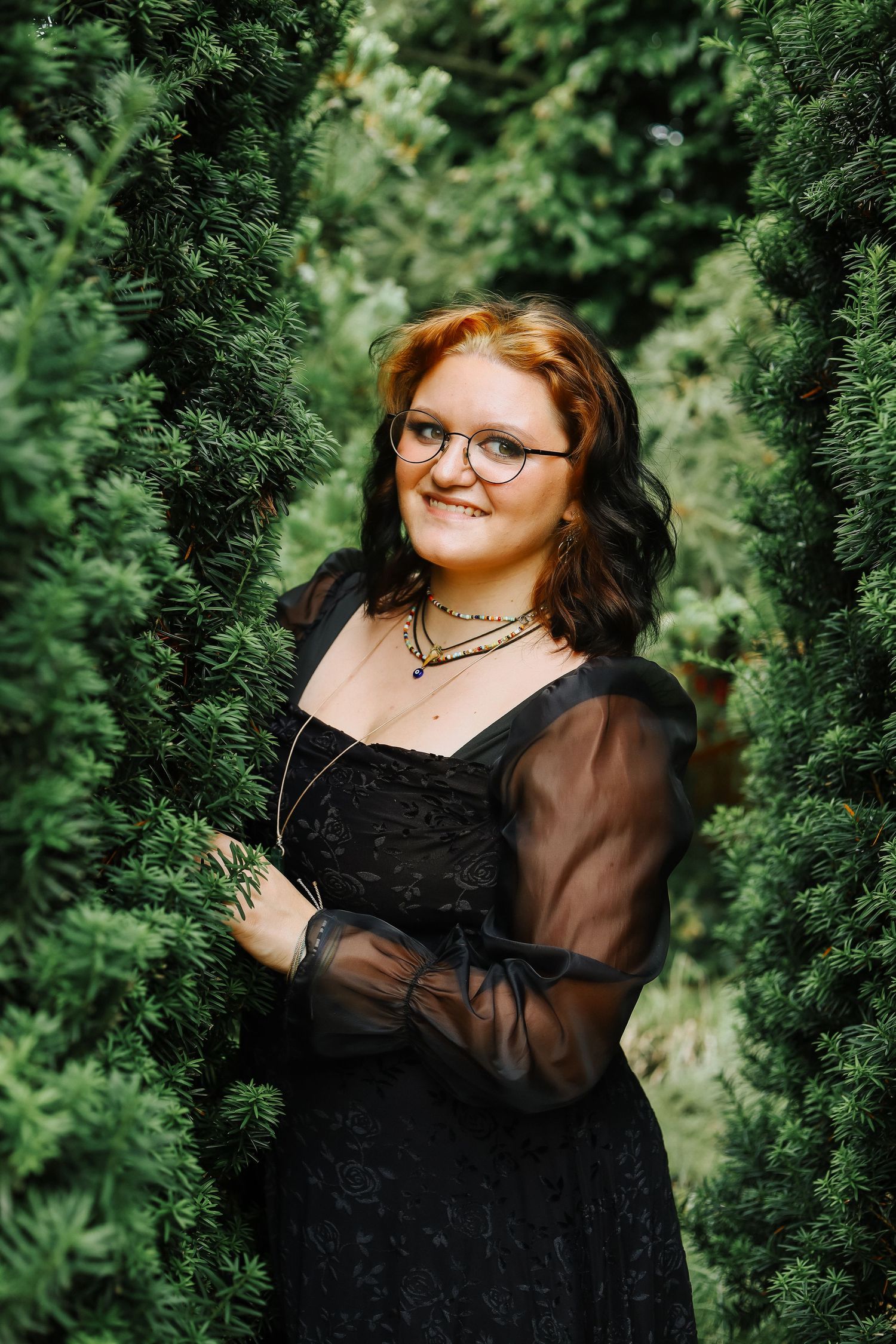 Person wearing black dress poses against evergreen trees in outdoor portrait session.