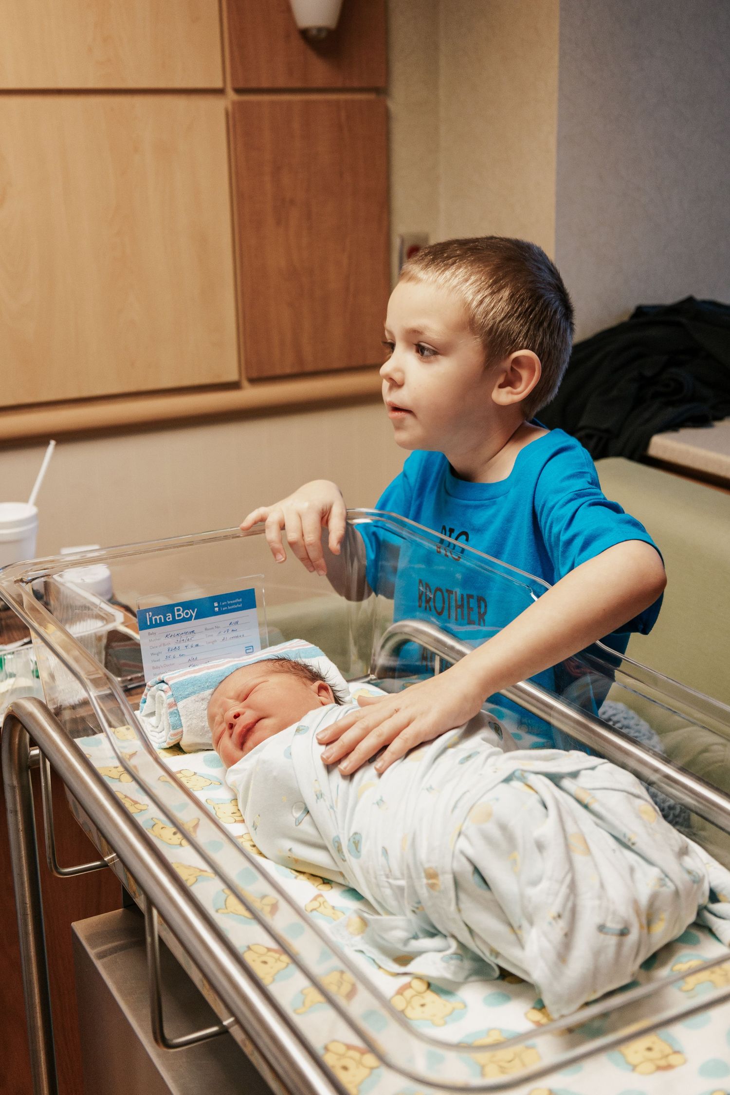 Young child in blue shirt looking at newborn baby in hospital bassinet.