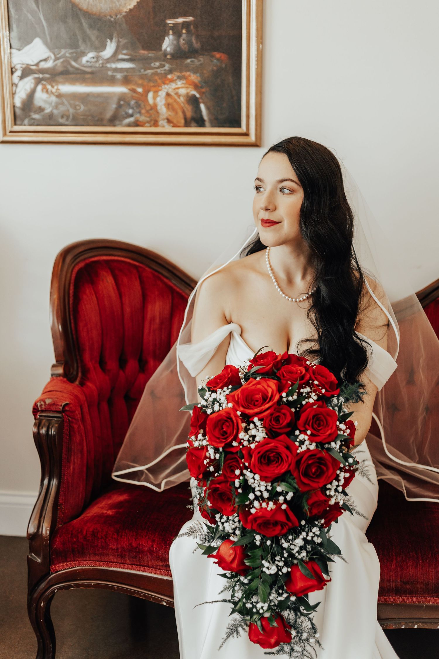 Elegant bridal portrait featuring cascading red rose bouquet on vintage velvet chair.