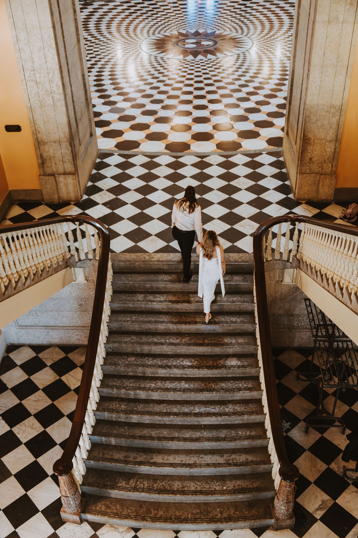 Two women ascending grand marble stairs in an elegant hall with striking black and white checkered floor pattern.