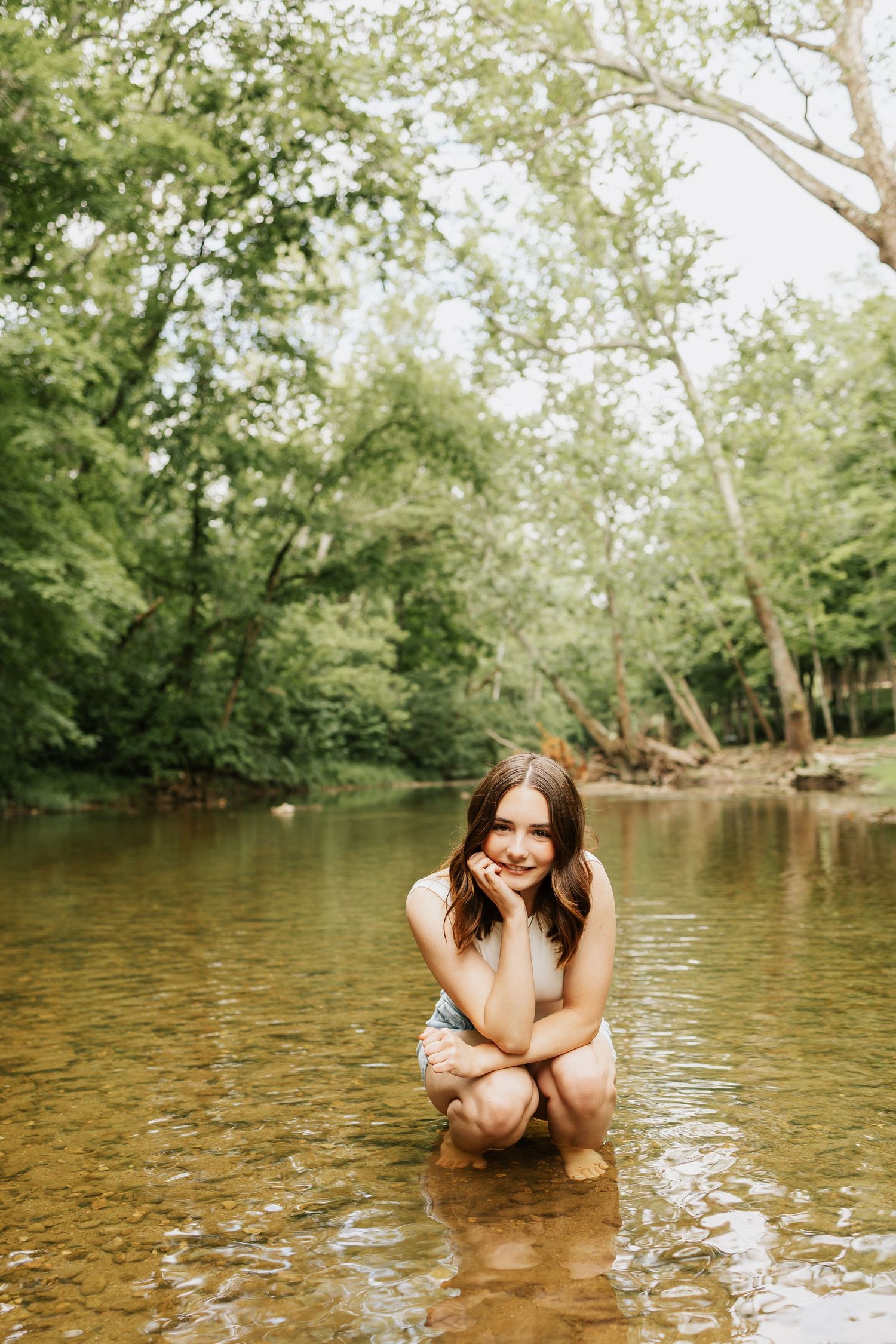 Young woman crouching in a shallow forest creek, smiling, surrounded by lush green trees in a serene outdoor setting.