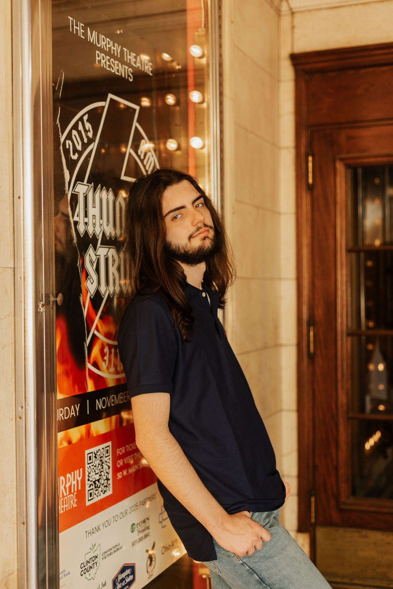 Person in navy polo leans against wall next to illuminated event poster.