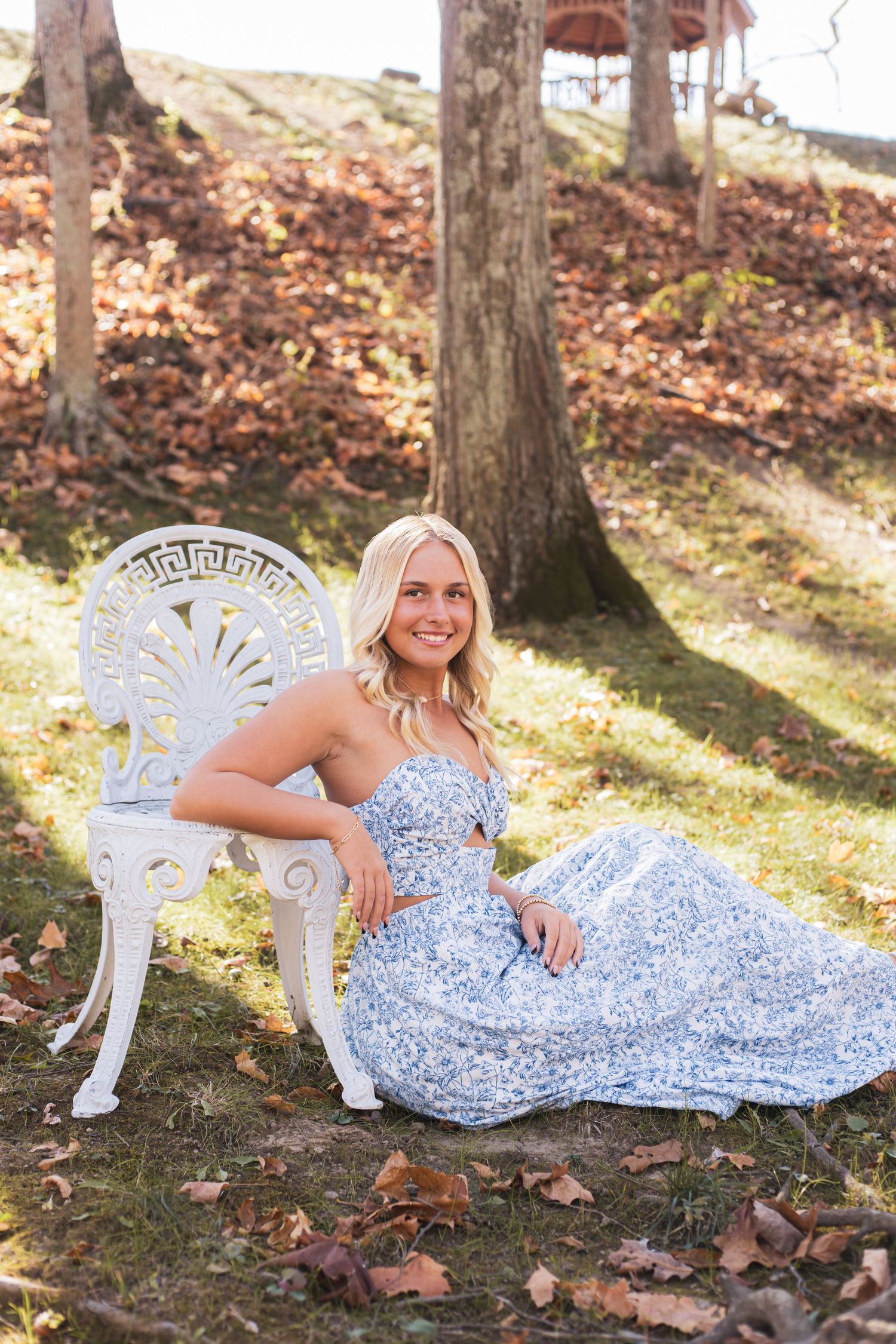 A light blue dress flows elegantly against fallen leaves and decorative white garden furniture.