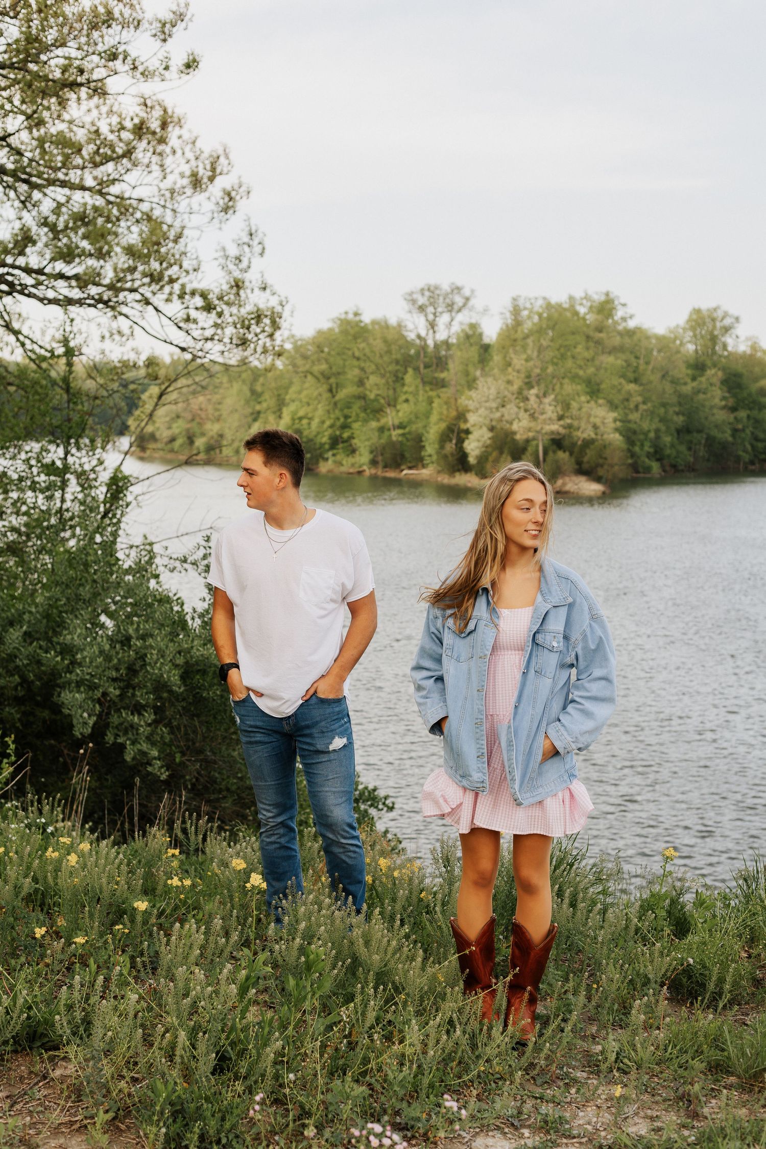 Couple standing by a serene lake at sunset wearing casual outfits in a natural outdoor setting.