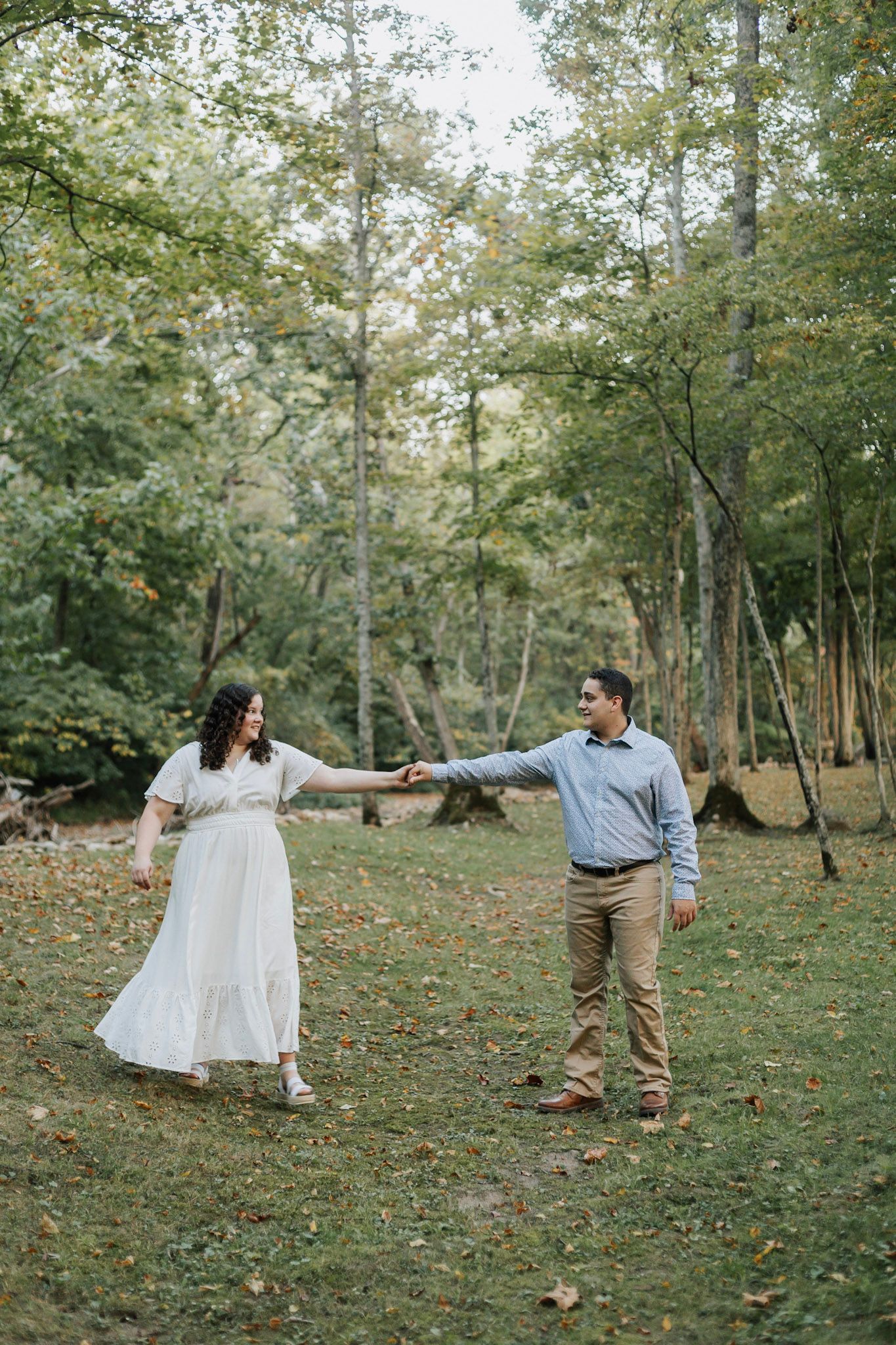 A couple dances playfully in a wooded area with fallen autumn leaves on the ground.