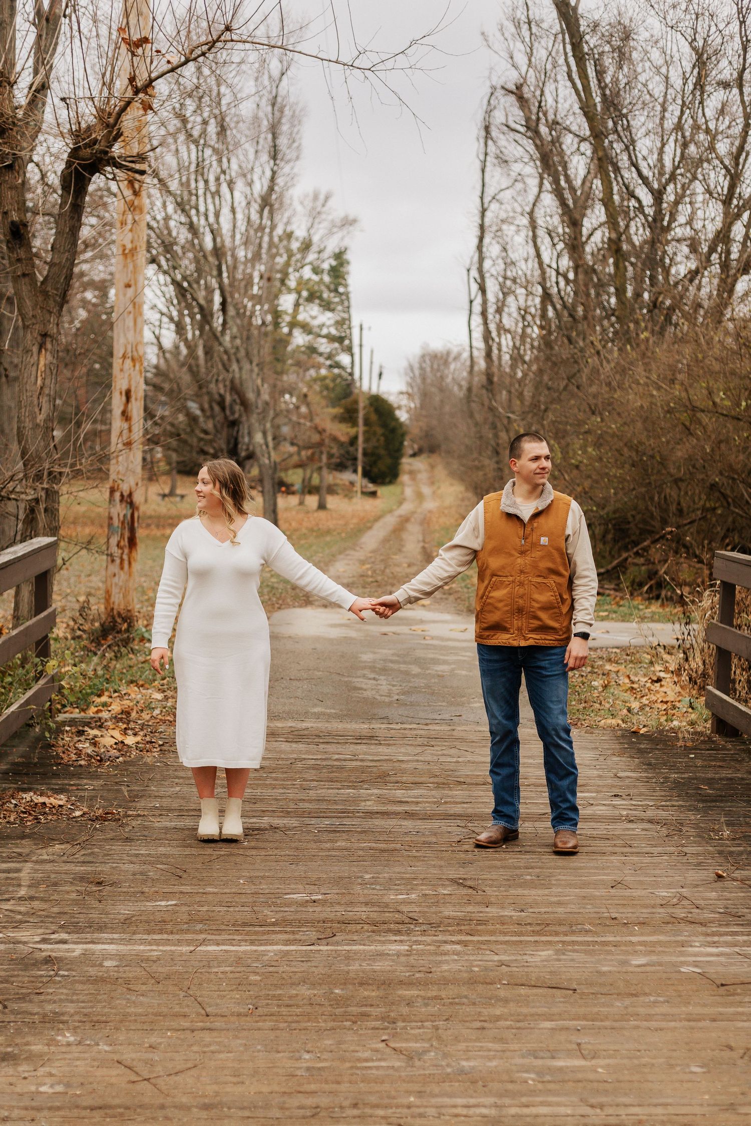 Couple walking hand in hand on wooden boardwalk surrounded by bare trees.