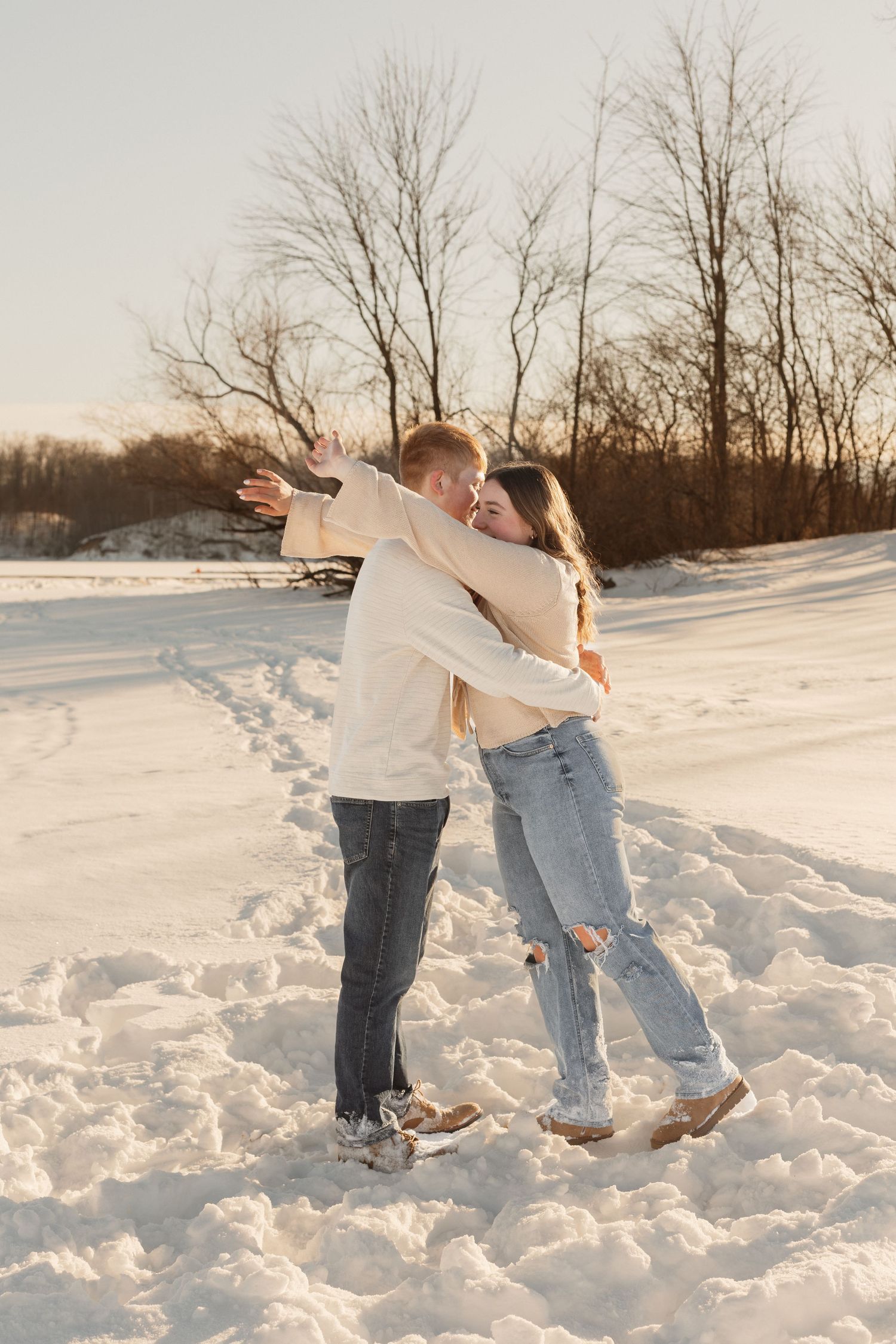 A couple dances playfully together in the snow at sunset with bare trees in the background.