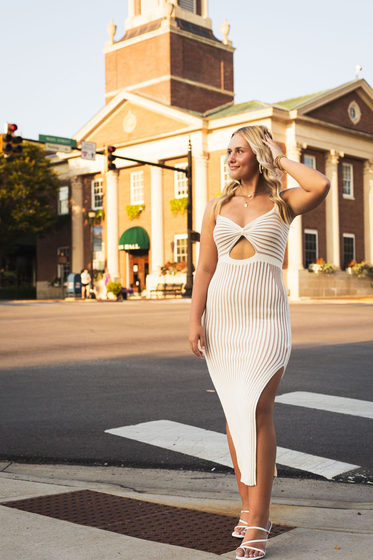 A person in a white striped dress poses on a street corner near brick buildings at sunset.