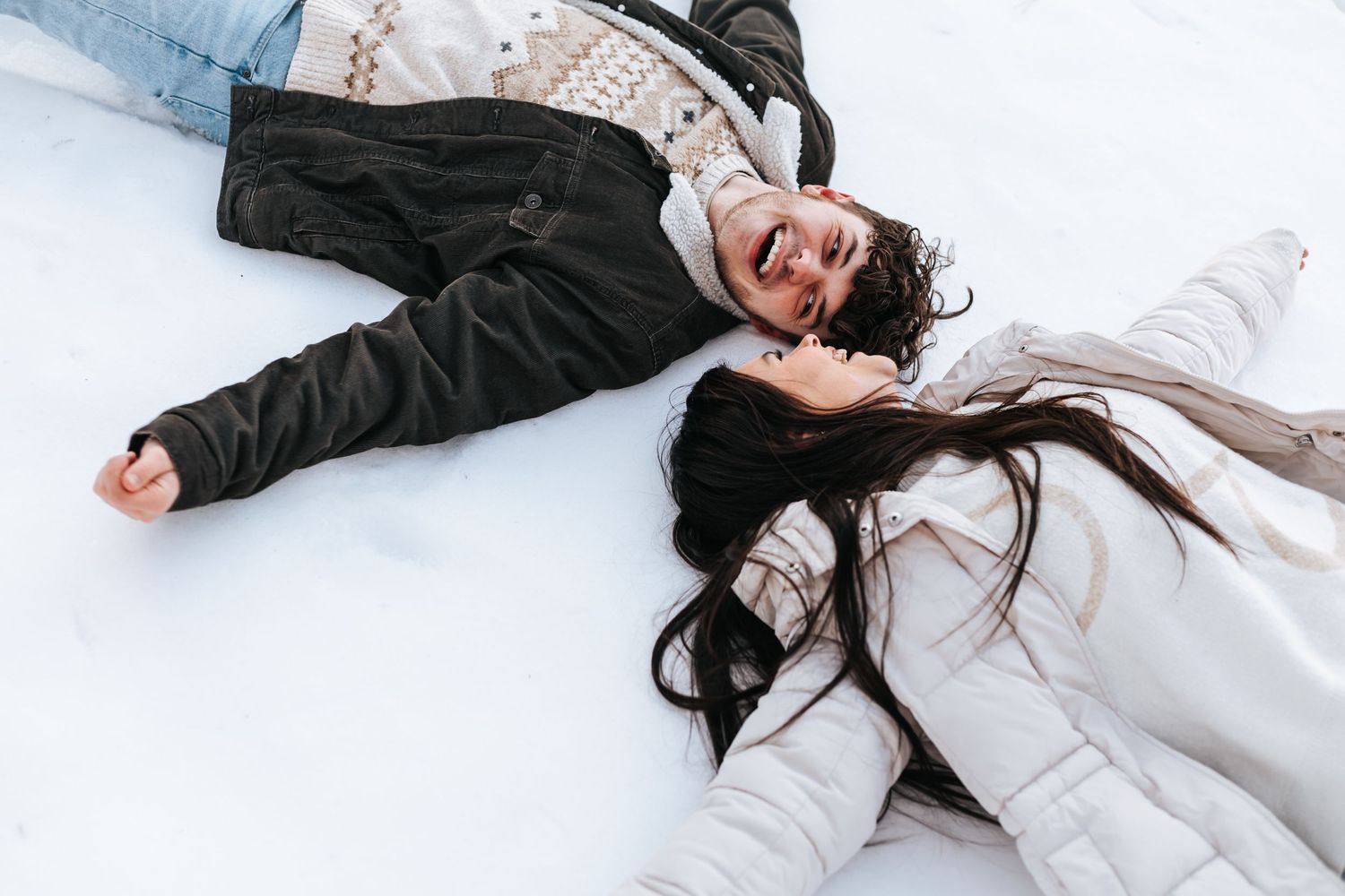 Couple lying in snow making snow angels while wearing winter coats.