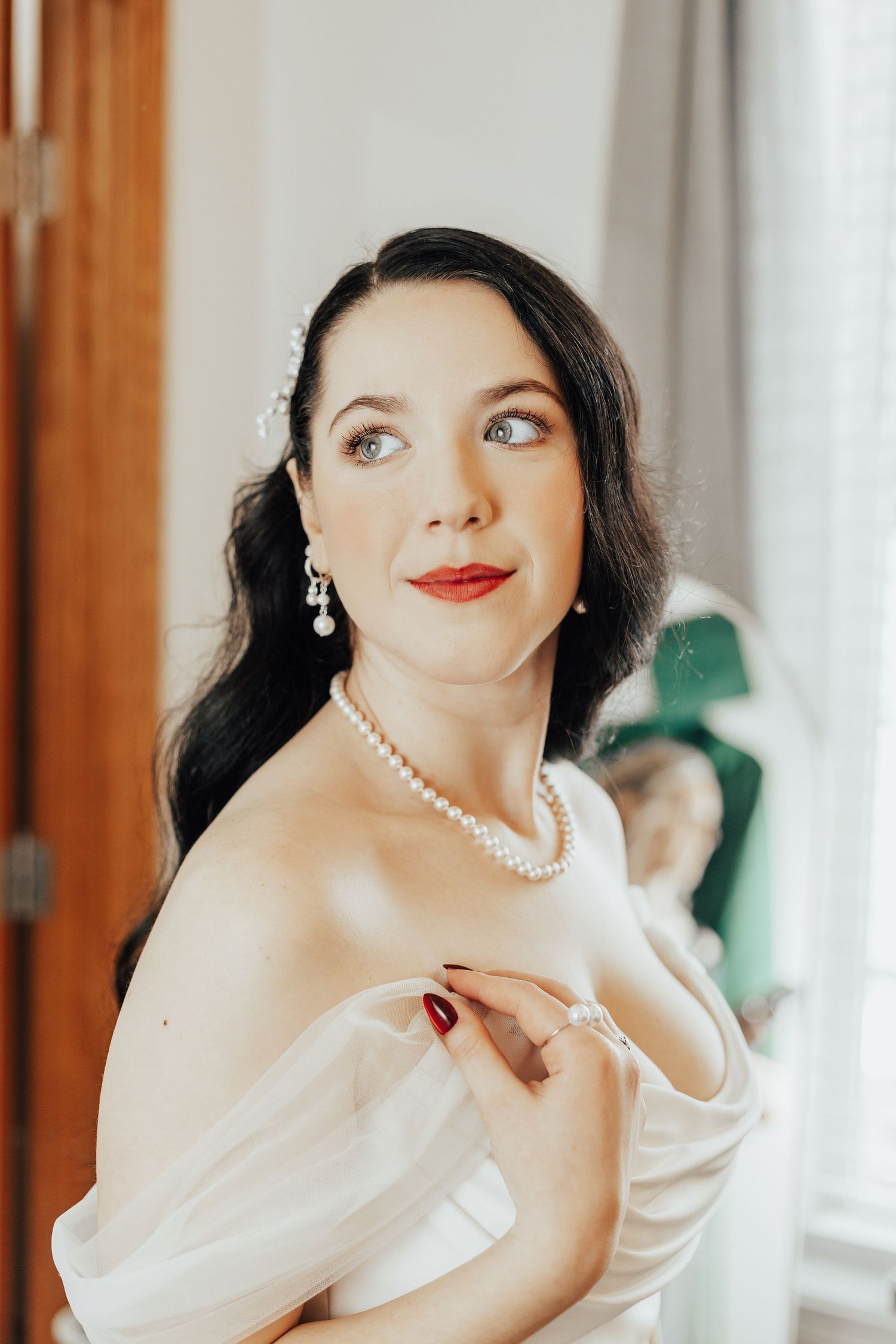 Elegant bride in white off-shoulder gown with pearl necklace and earrings, dark hair, red lips, gazing sideways.