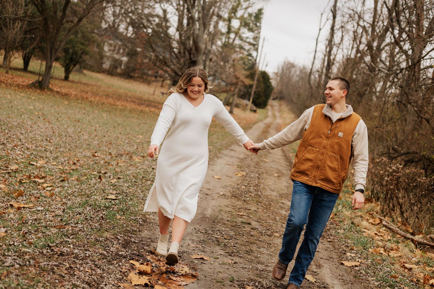 Couple running playfully on dirt path surrounded by autumn trees.