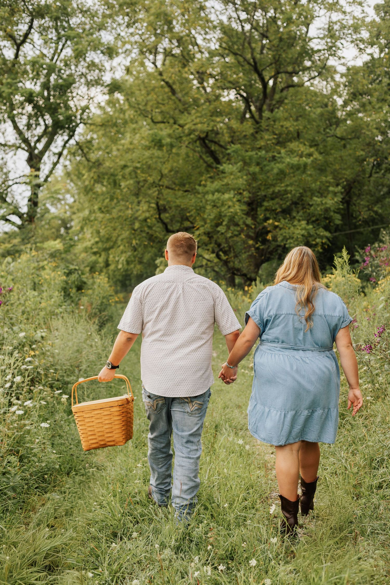 Couple walking through wildflower meadow holding hands and carrying picnic basket.