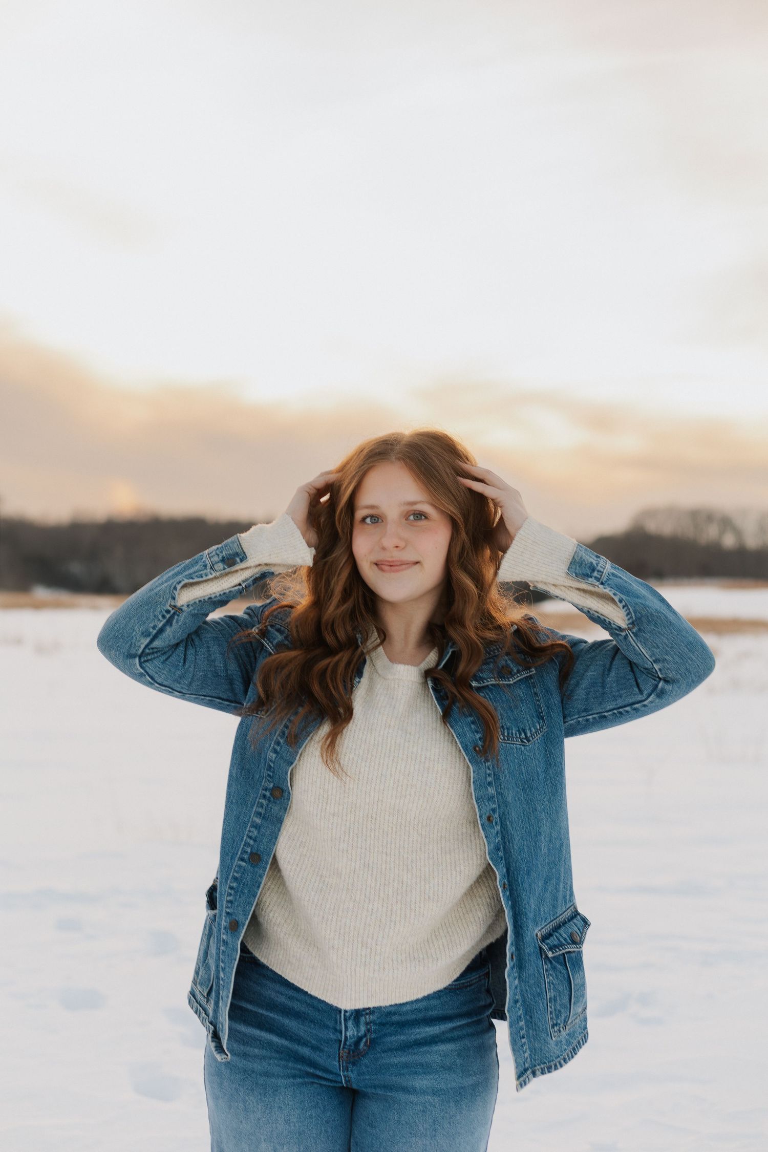 Person in denim jacket and jeans poses against snowy winter landscape at sunset.