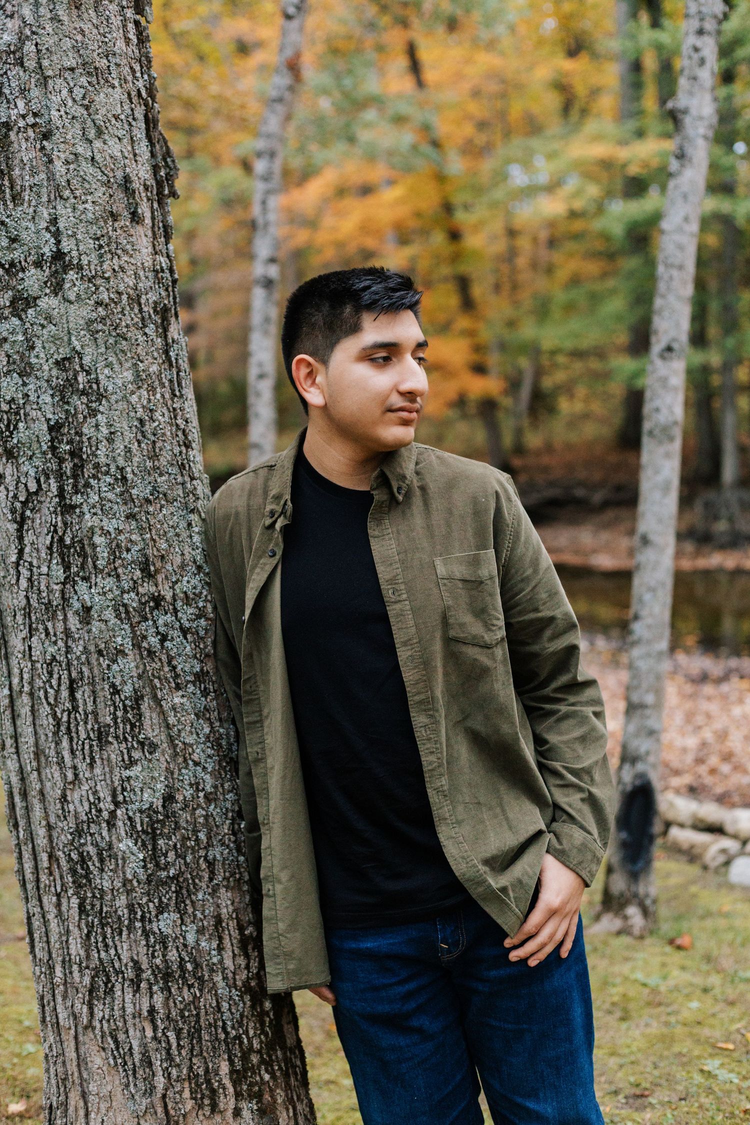 Person in olive jacket and jeans leaning against large tree trunk in fall forest.