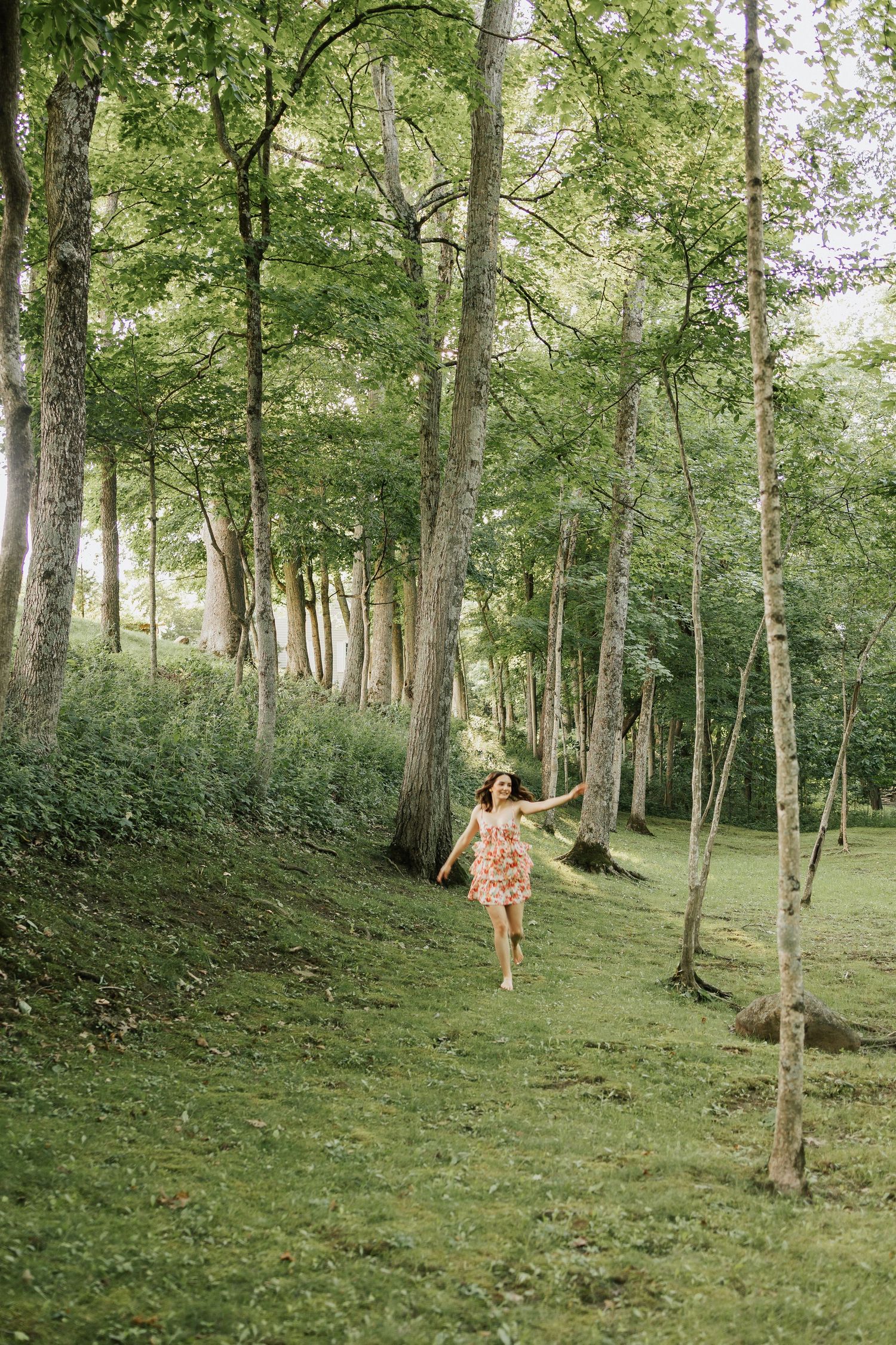 A woman in a floral dress walks through a lush green forest path lined with tall trees on a sunny day.