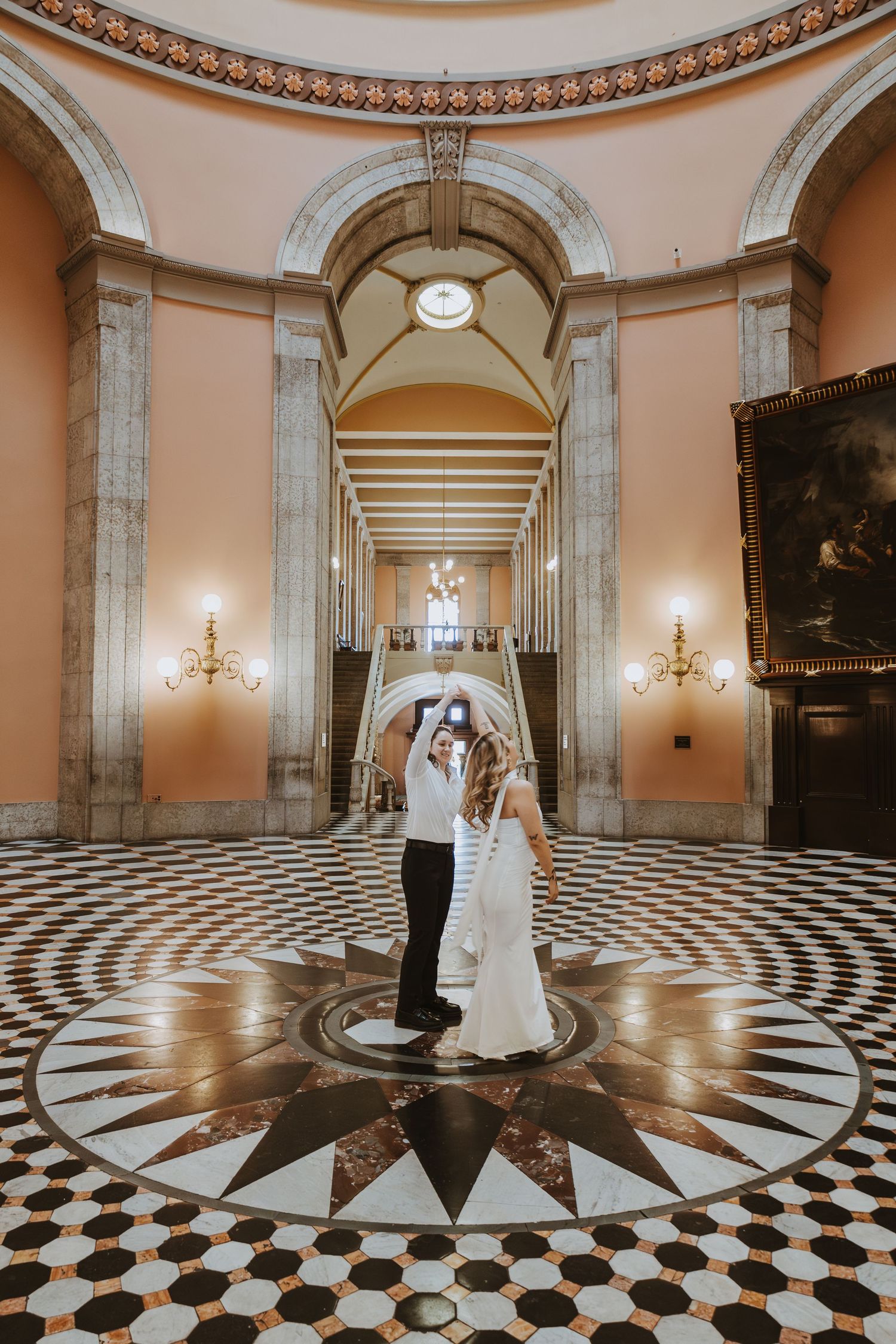 Bride and groom dancing on a black and white geometric marble floor inside an elegant domed hall with arched columns.