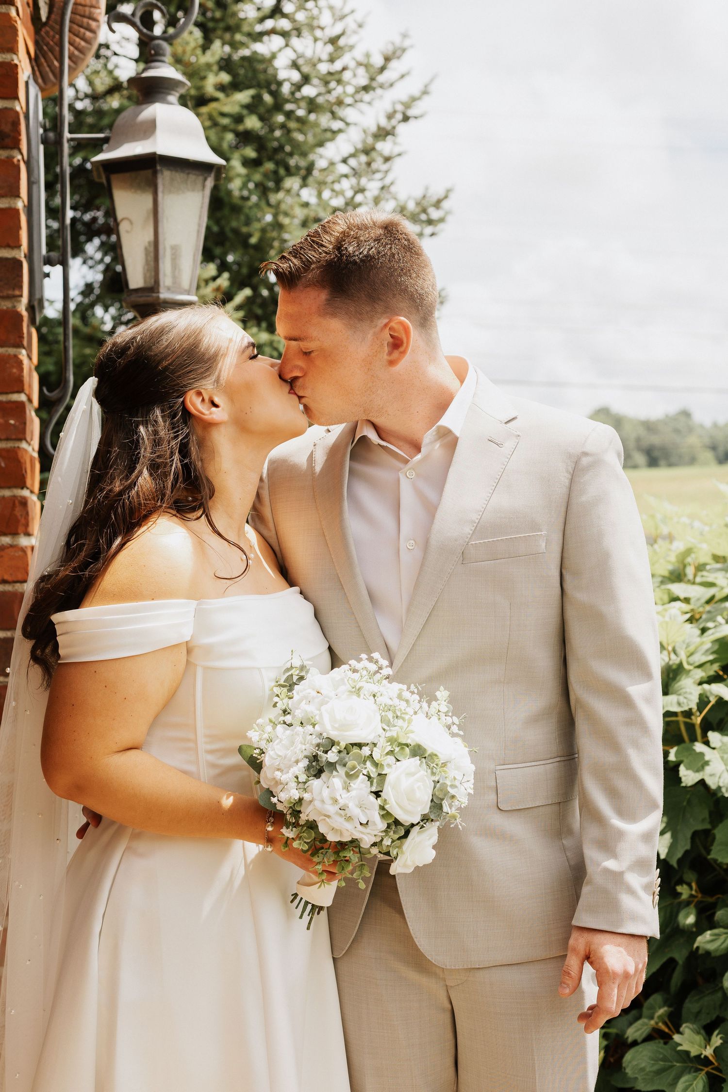 Romantic wedding moment captured in front of brick wall with white floral bouquet.