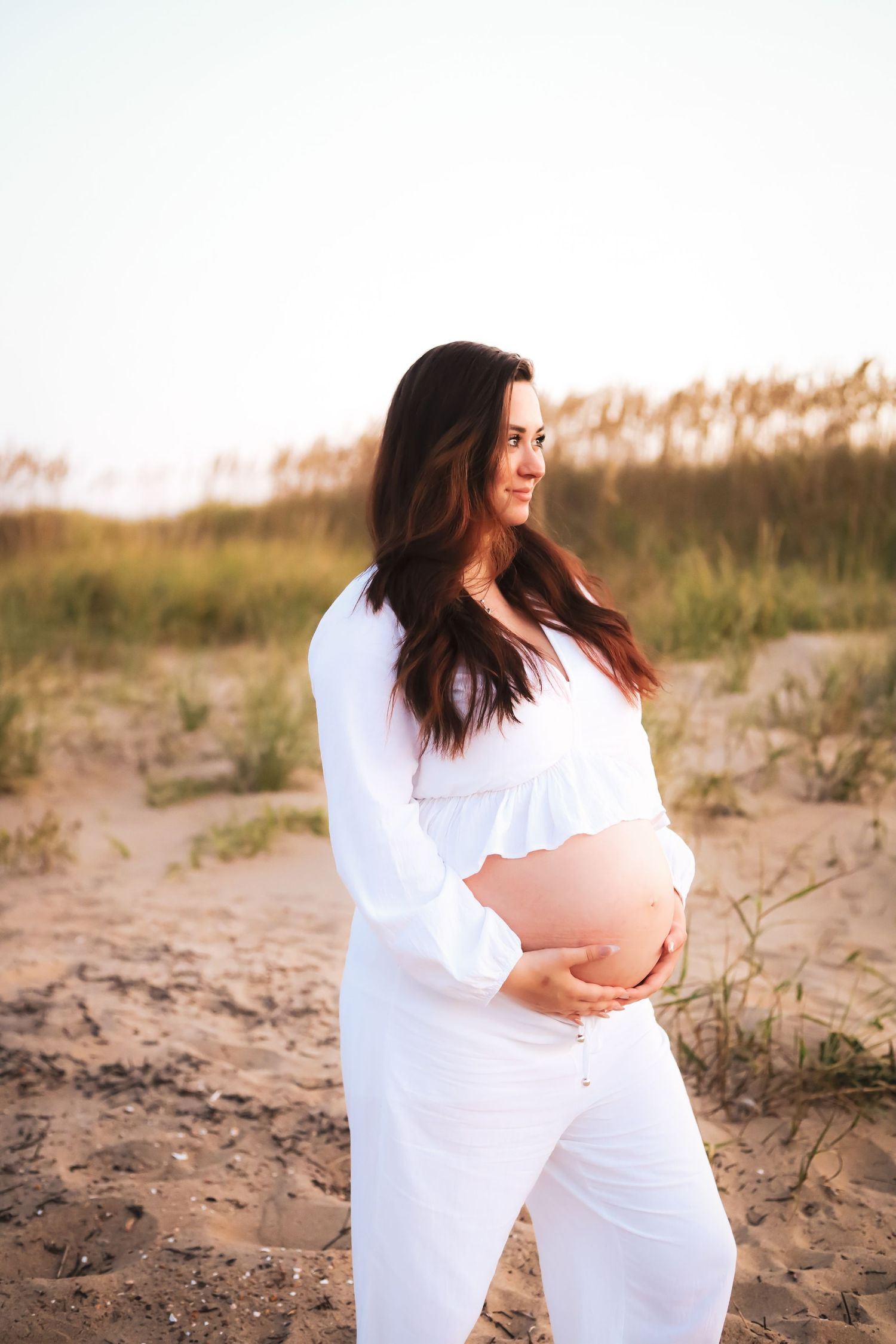 Maternity photoshoot on sandy beach dunes at sunset with flowing white fabric.