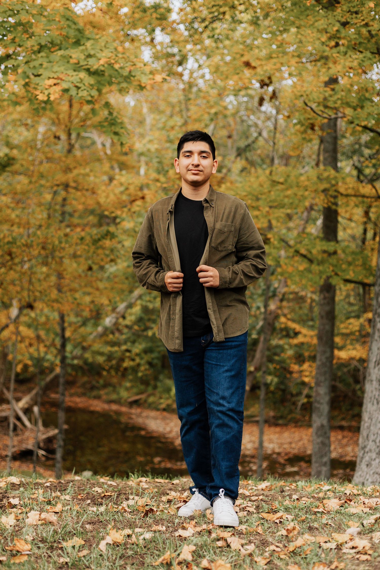 Person standing on fallen leaves wearing olive jacket and jeans in autumn forest.