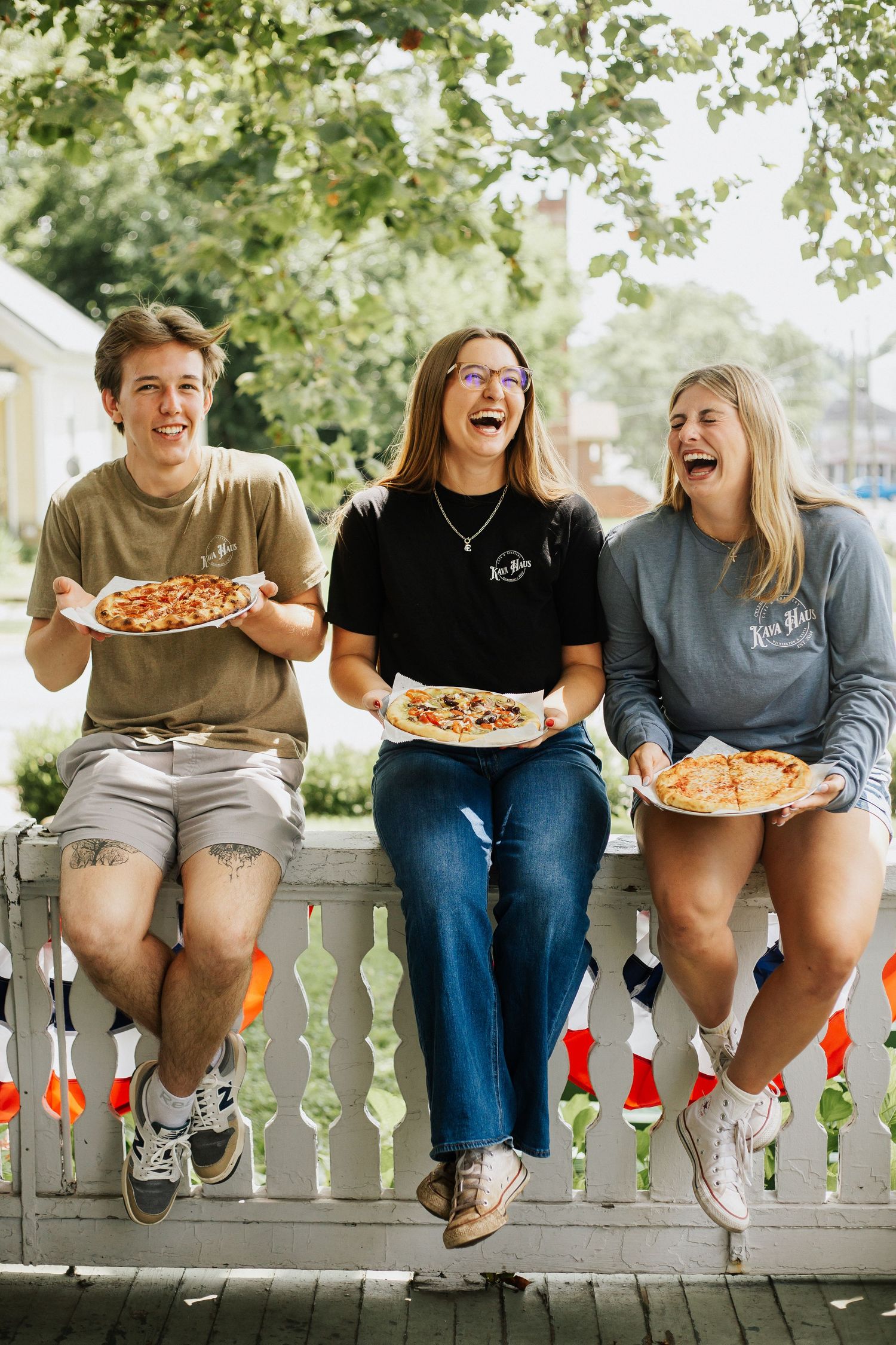 Friends sitting on a white railing outdoors sharing pizza and laughing together.