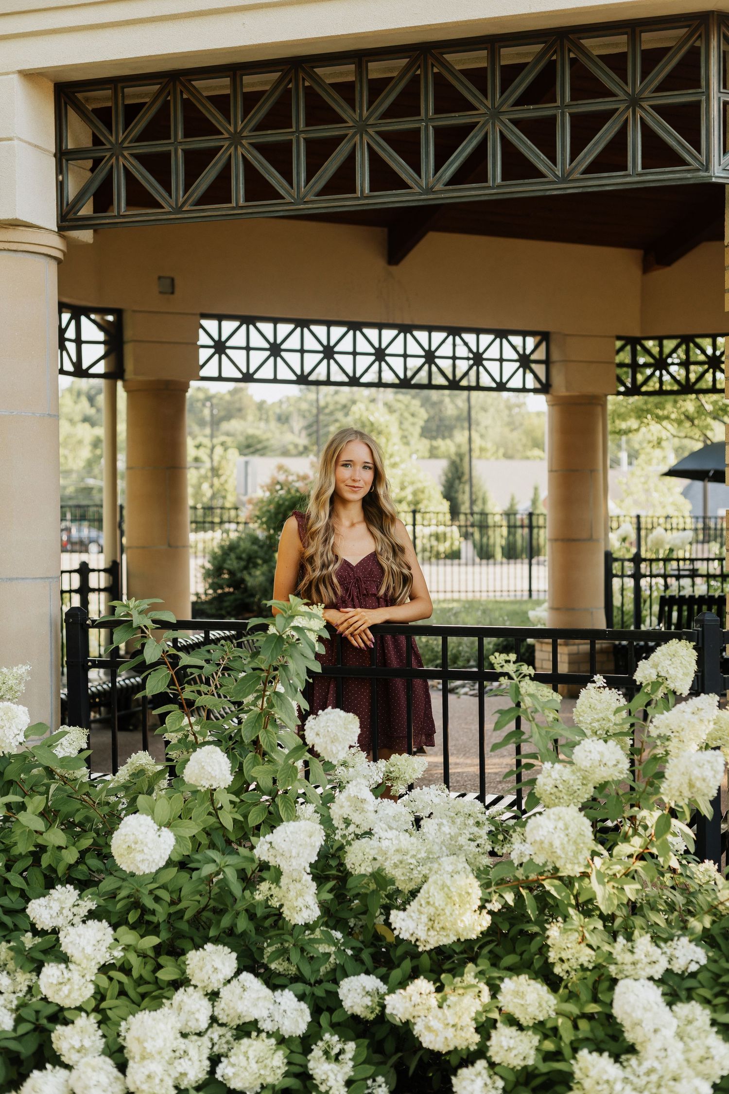 Young woman in burgundy dress poses behind white hydrangeas at an elegant outdoor pavilion.