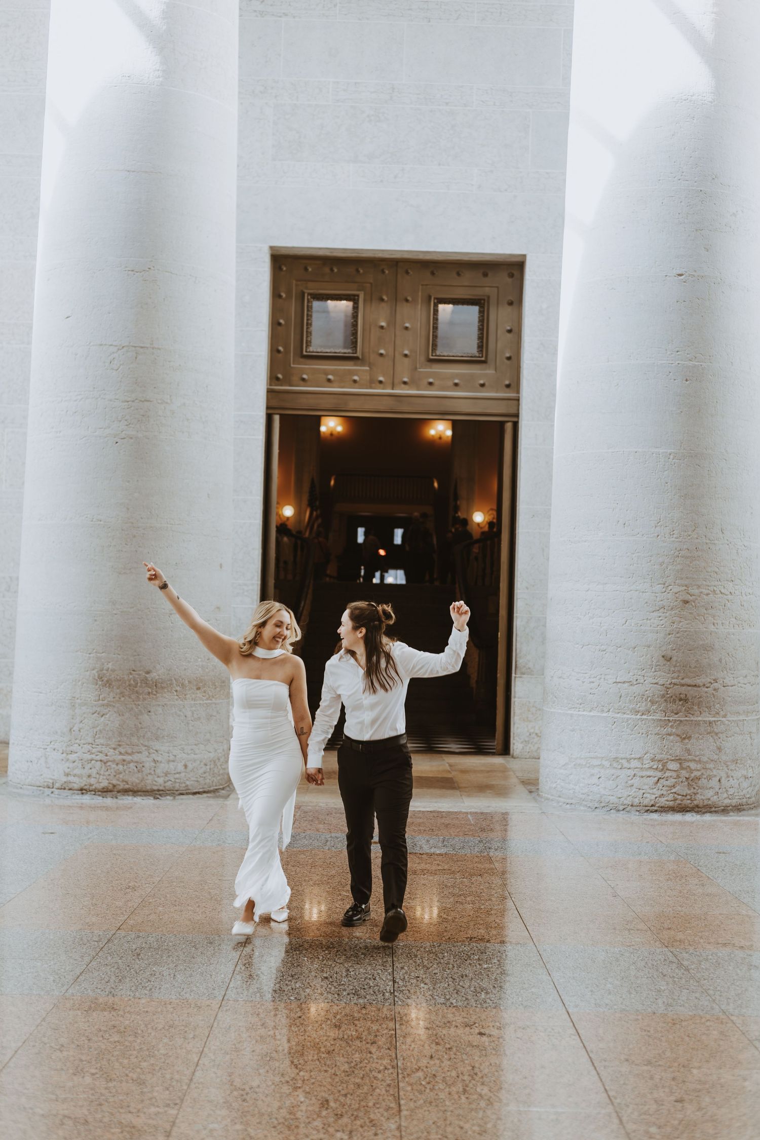 Two brides celebrate joyfully holding hands, walking between grand white columns at an elegant wedding venue.