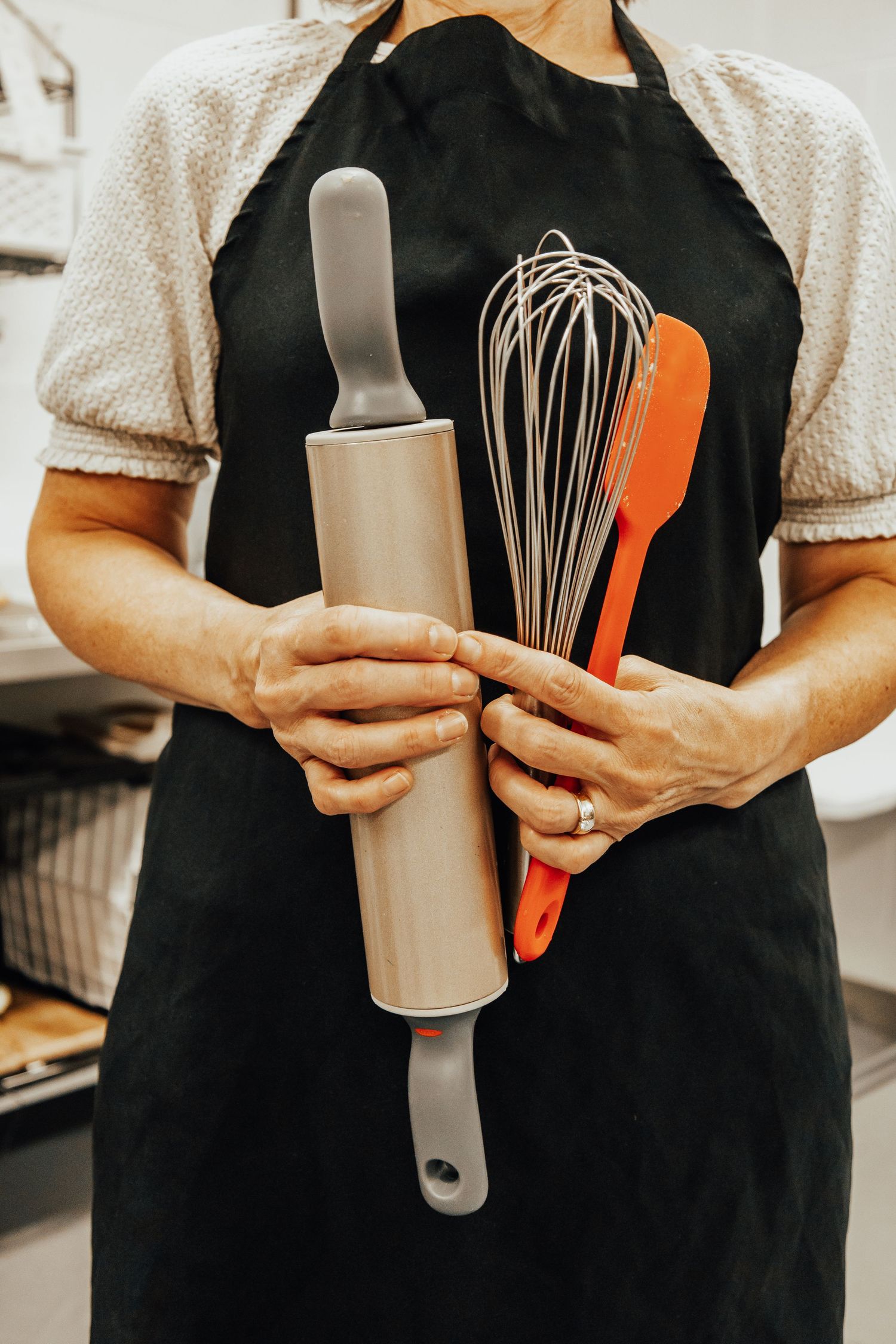 Close-up of kitchen tools including whisk and spatula being held against black apron.