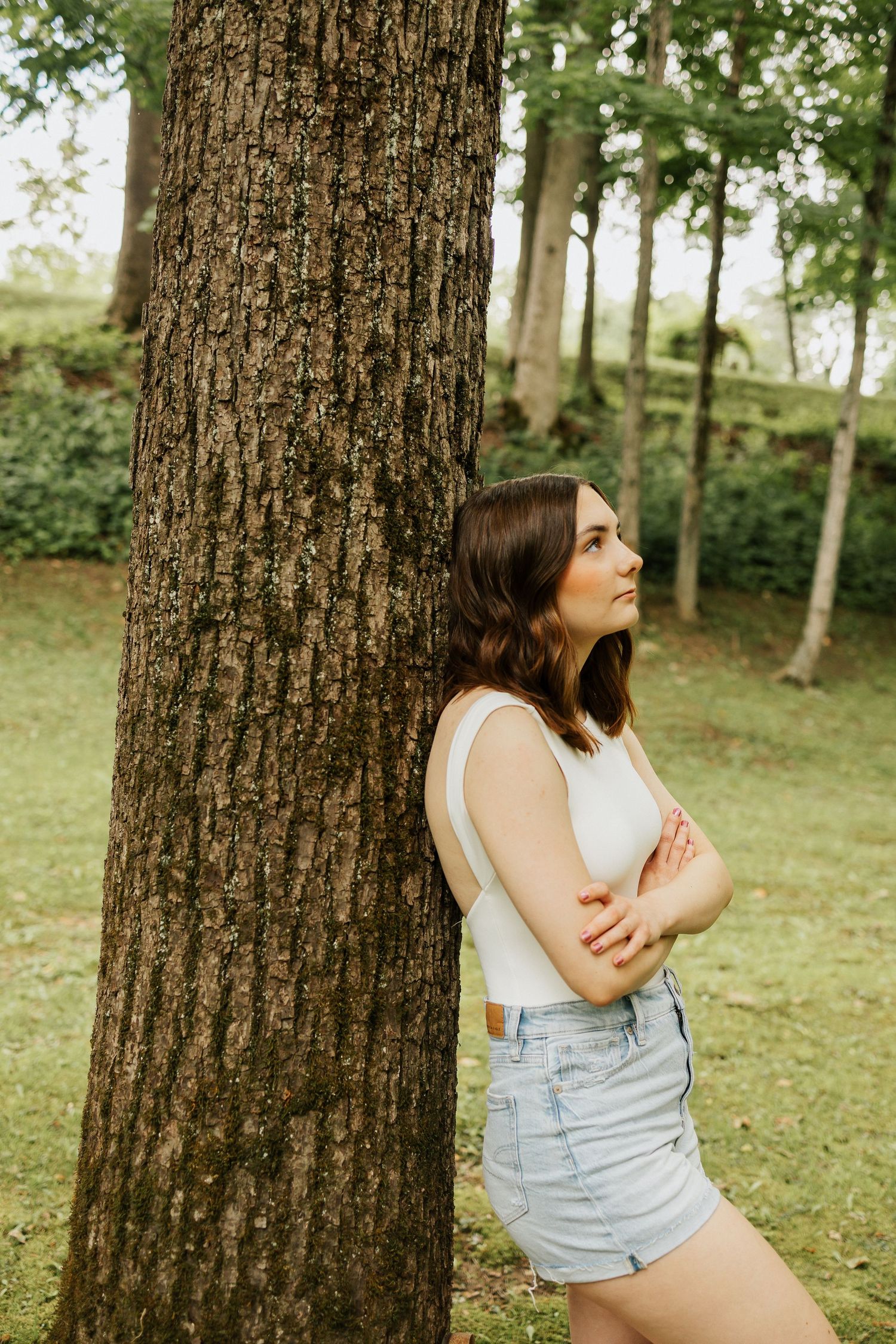 Young woman in white top and denim shorts leaning against a large tree trunk in a lush green park.
