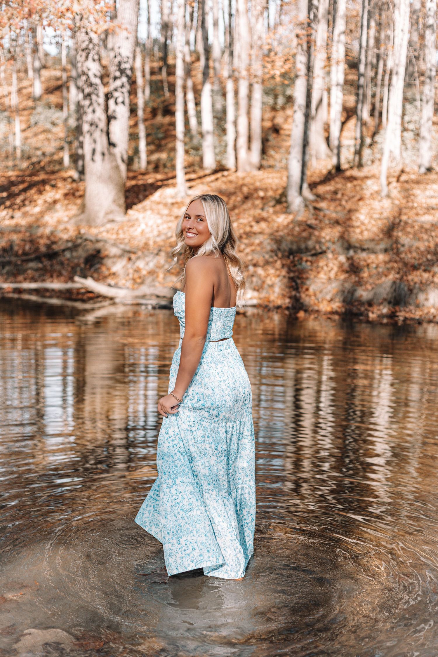 A flowing light blue dress swirls near a reflective pond in an autumn forest setting.