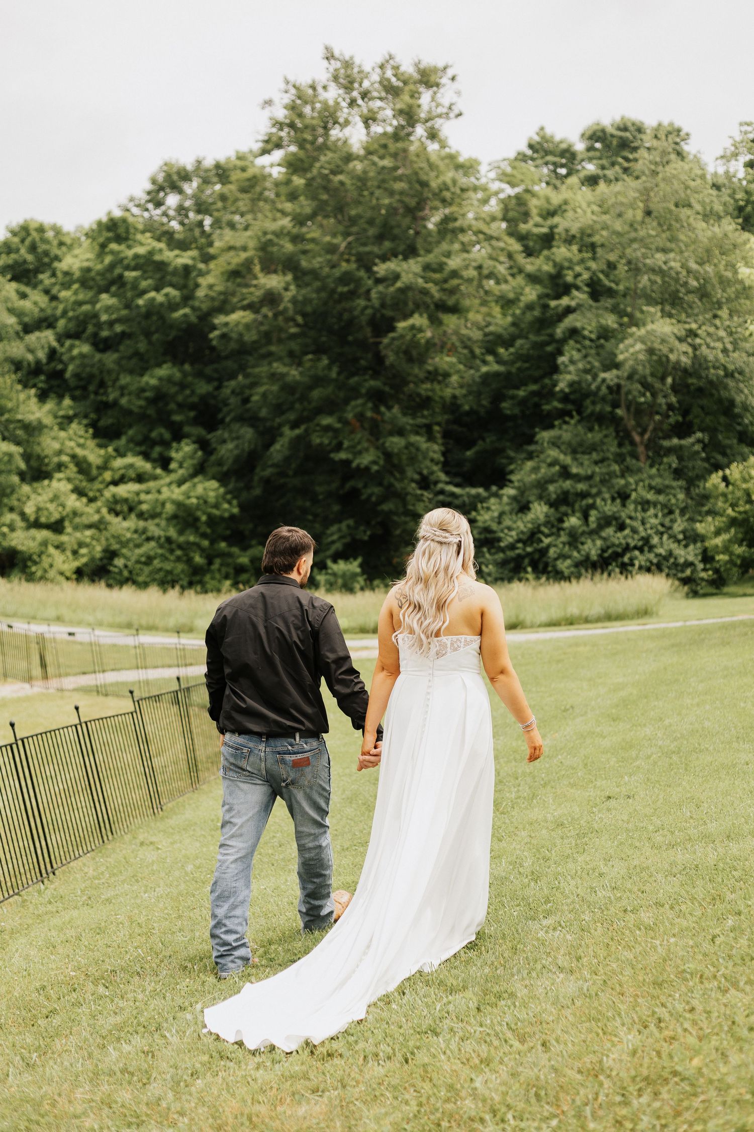 Couple walking hand in hand across a green field with flowing white dress trailing behind.