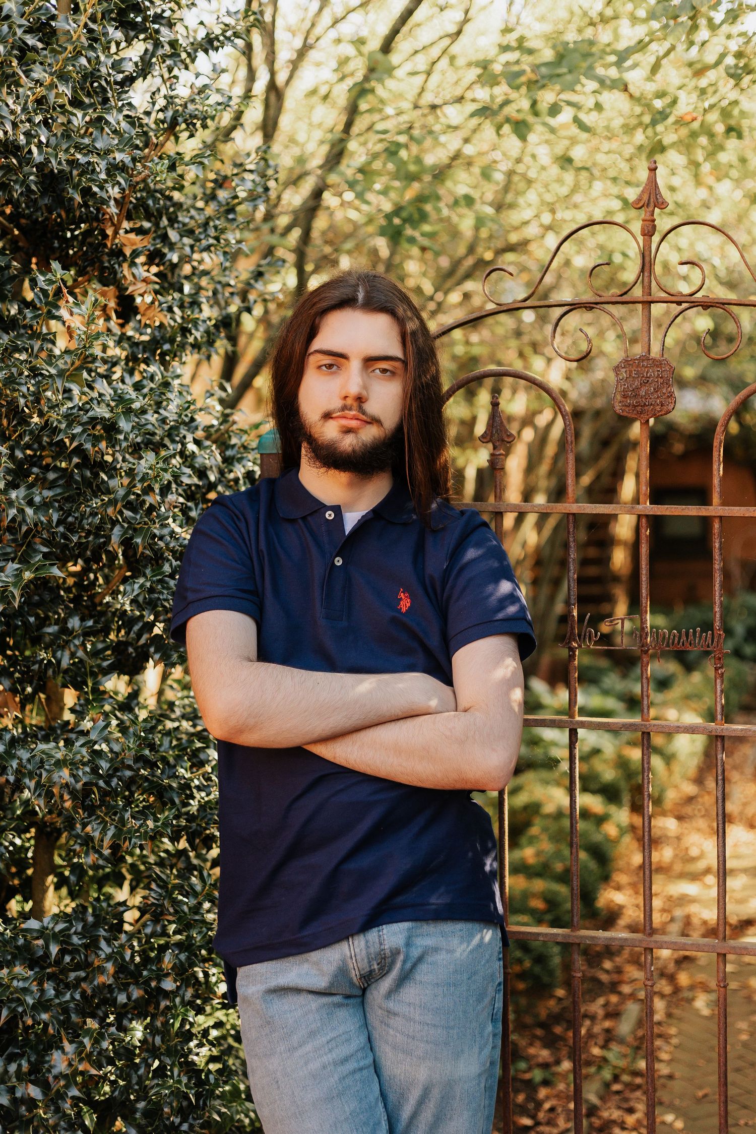 Person in navy polo stands by ornate iron gate surrounded by greenery.