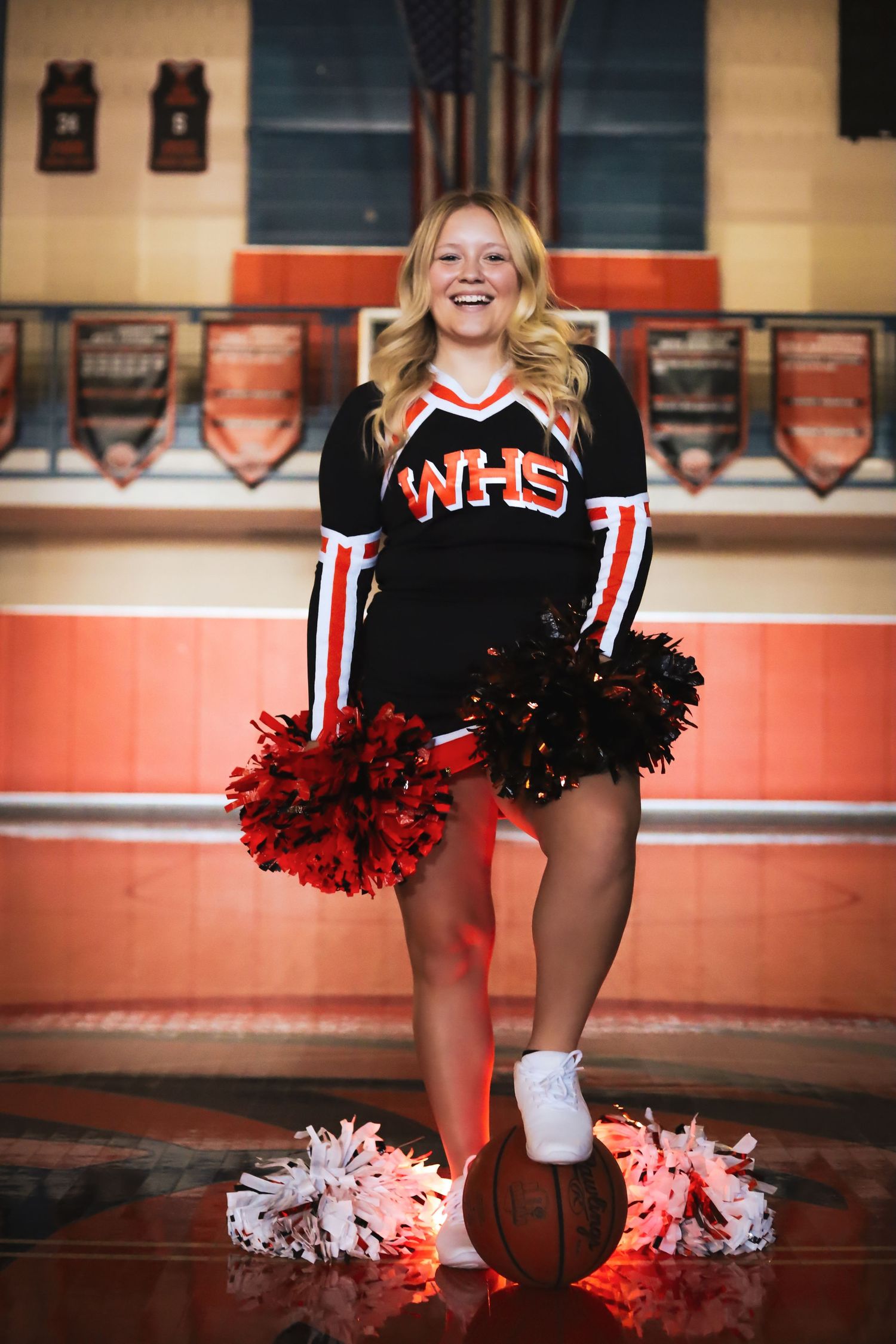 Cheerleader in black and red uniform with pom poms performing on a polished gym floor.