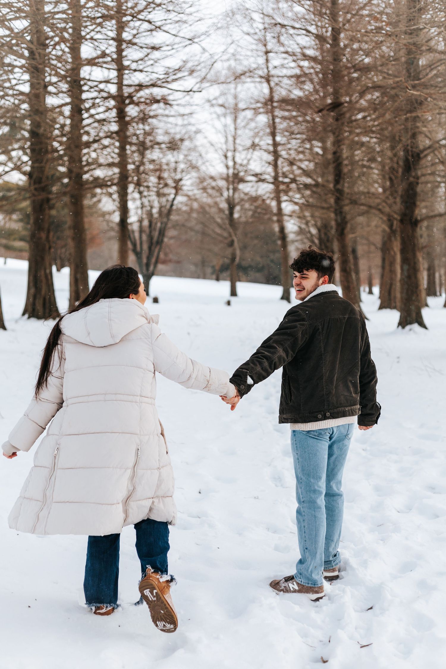 Couple holding hands while walking through snowy forest path in winter attire.