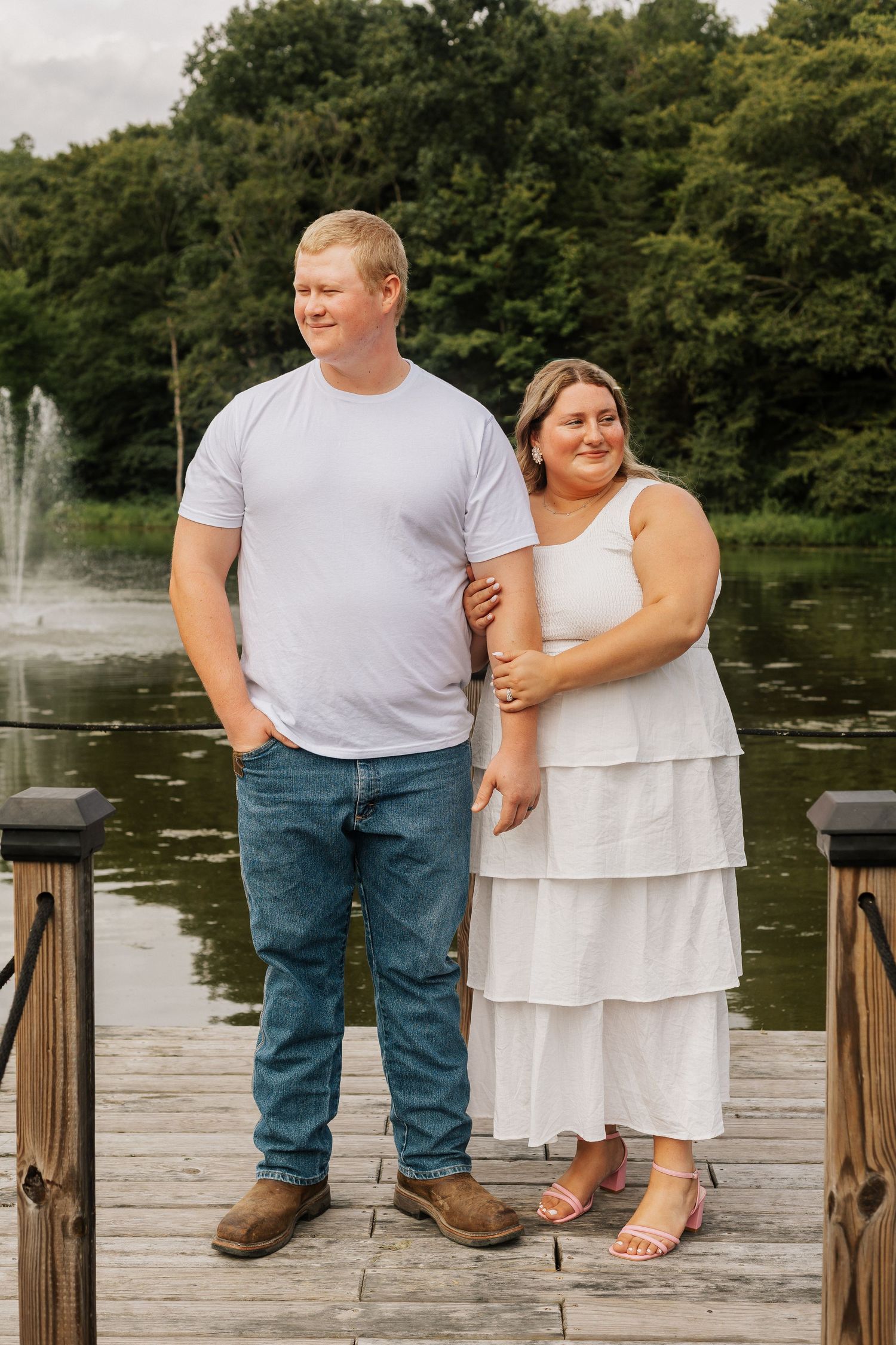 A couple stands together on a wooden dock with a fountain and pond in the background.