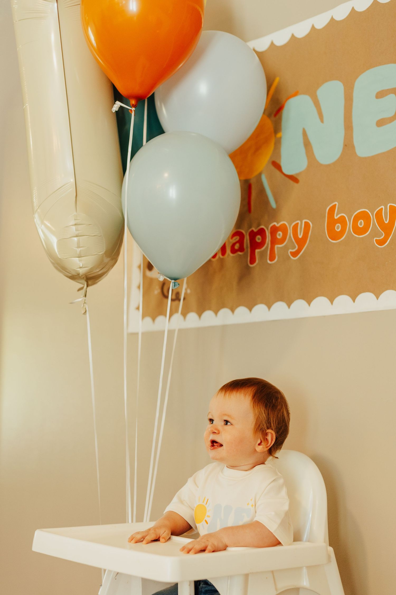 Baby sits in high chair with orange and white balloons at first birthday party celebration.