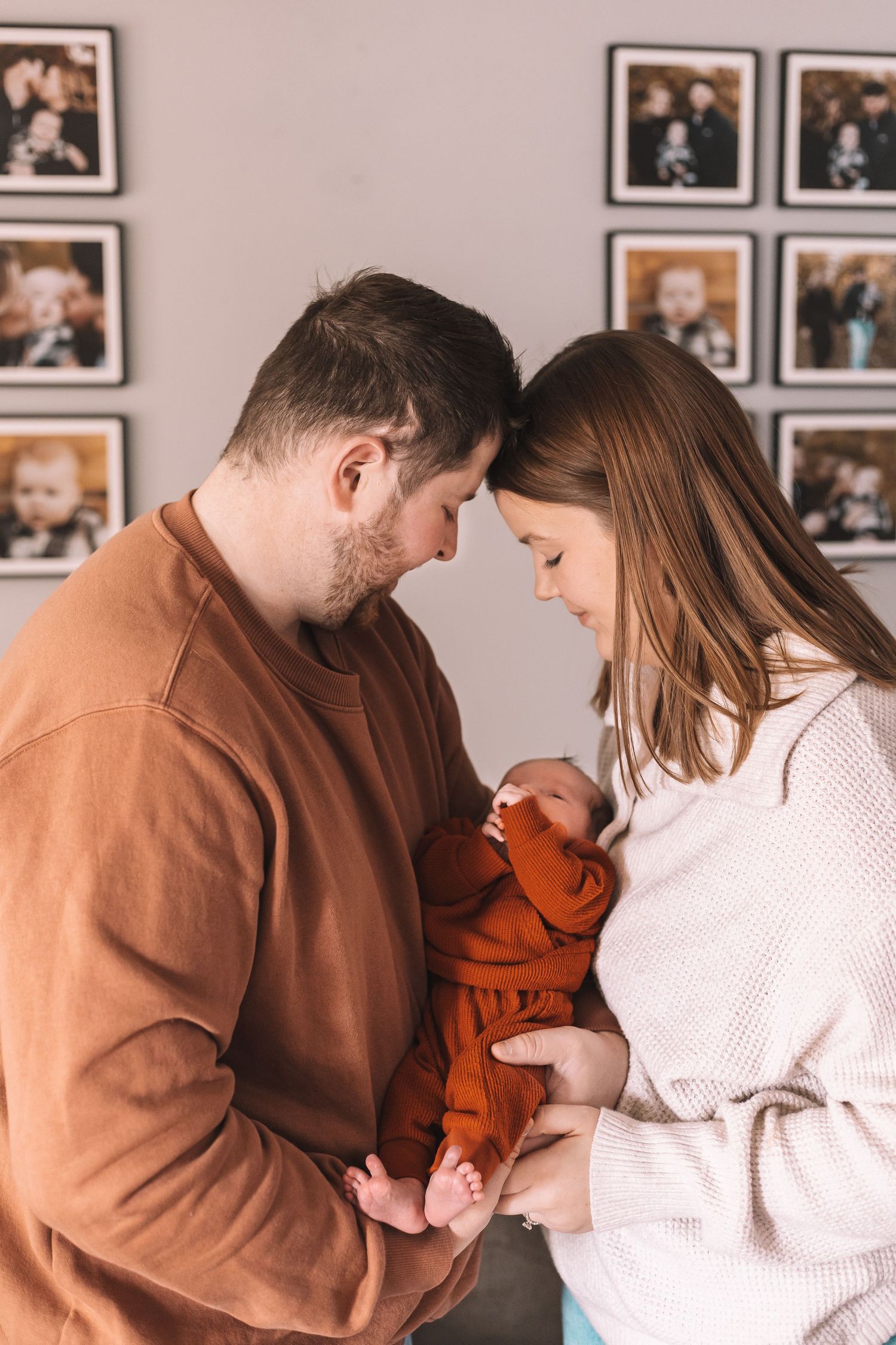 Parents sharing tender moments with their newborn in front of a wall of family photos.