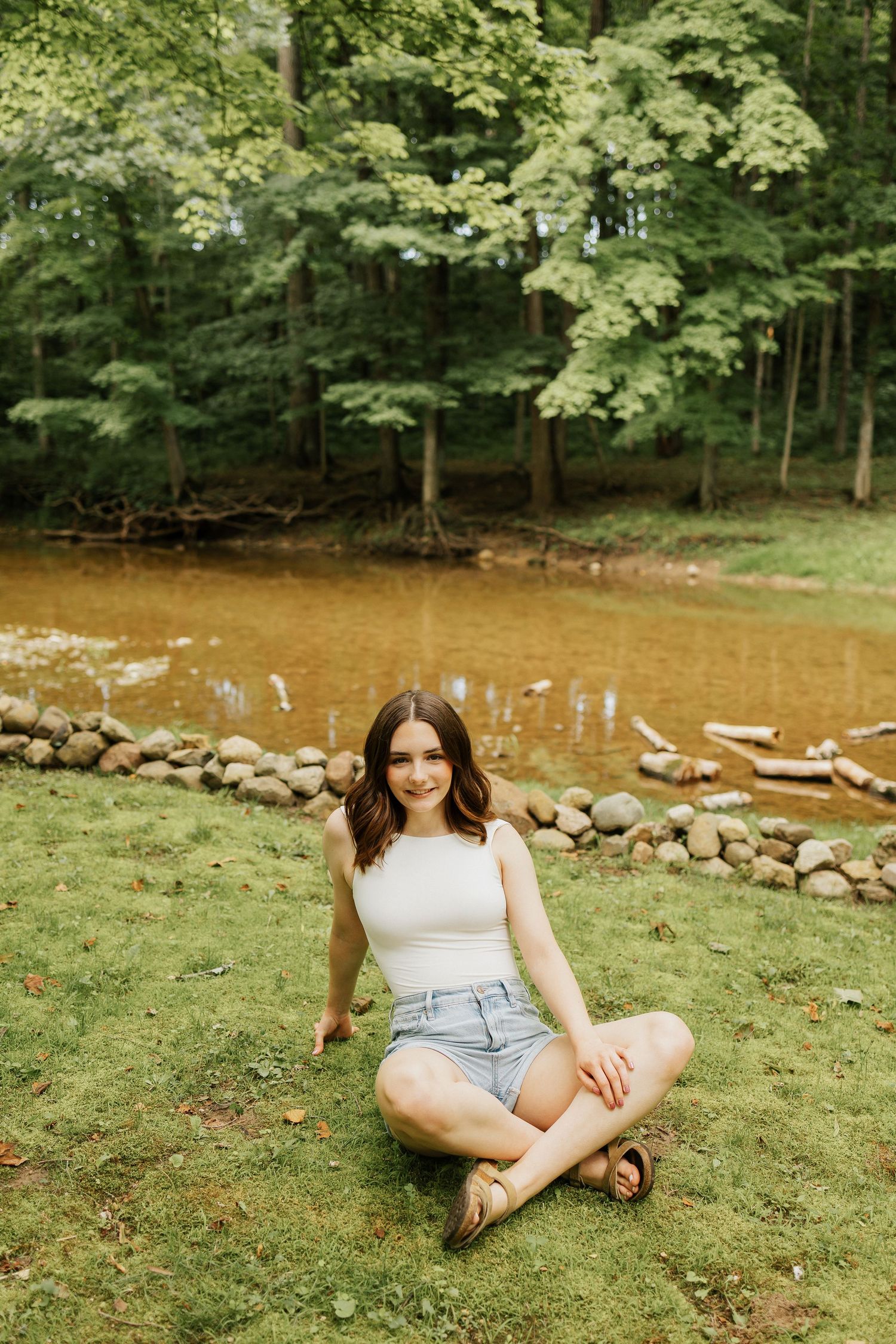 Young woman sitting cross-legged on grass near a rocky creek surrounded by lush green trees in a serene outdoor setting.