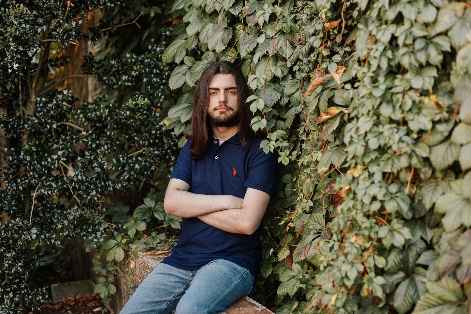 Person in navy polo sits against ivy-covered wall with arms crossed.