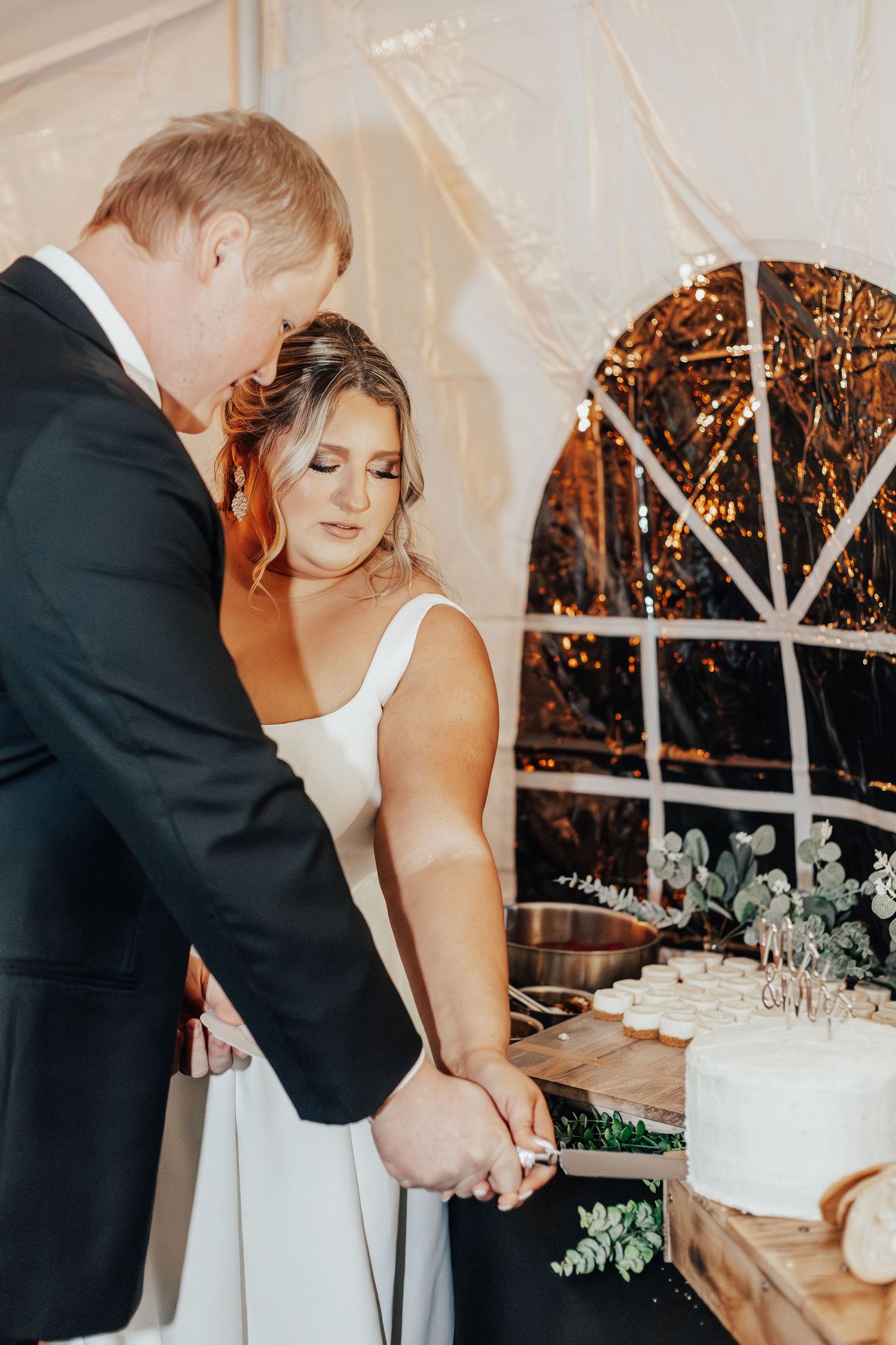 Couple cuts their elegant white wedding cake together at indoor reception.