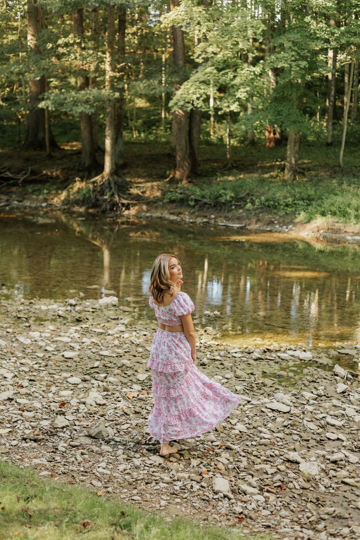 Woman in flowing pink dress walks along rocky riverbank in wooded setting.