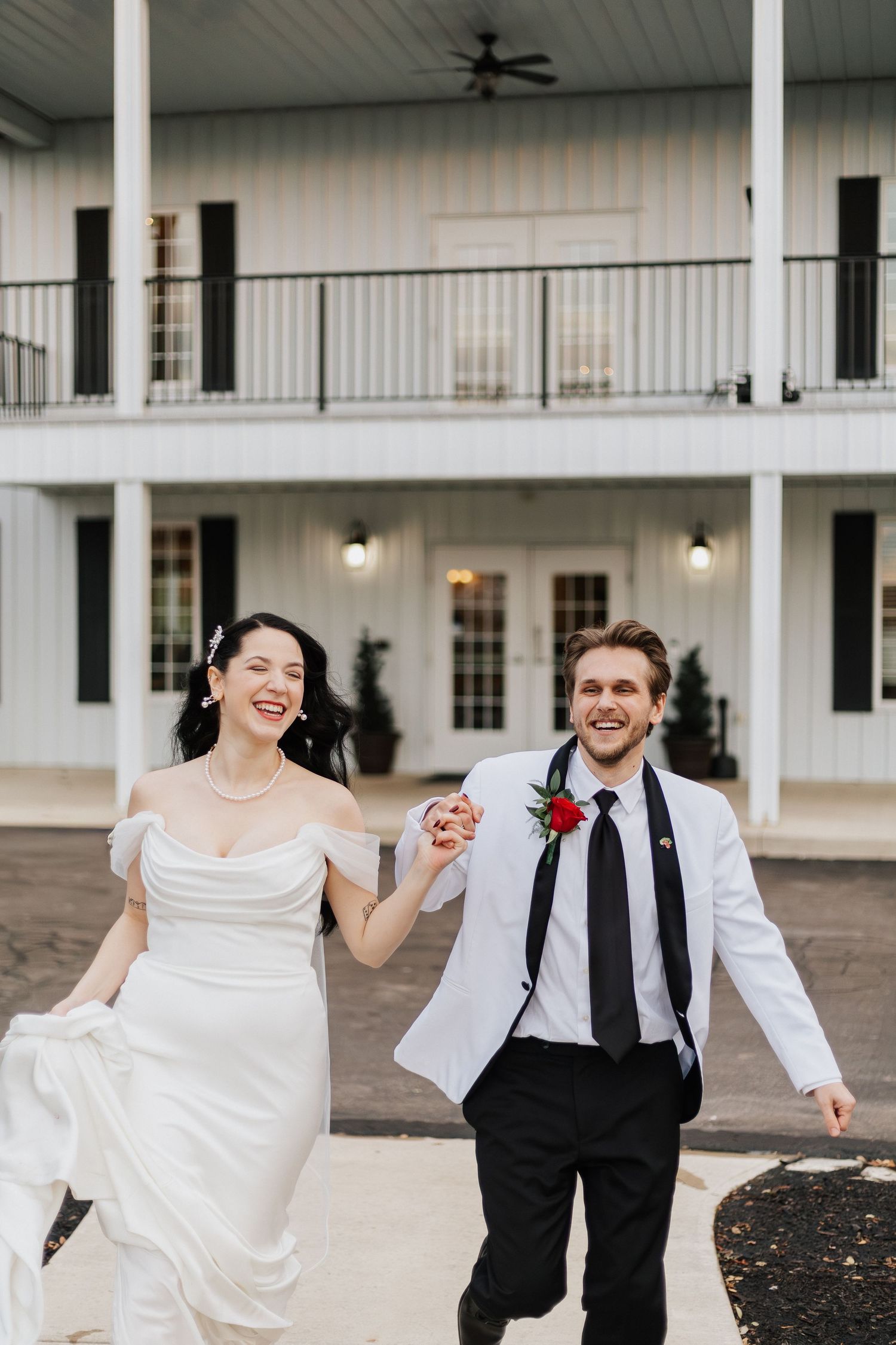 Joyful couple running and dancing together in formal wedding attire against white building exterior.