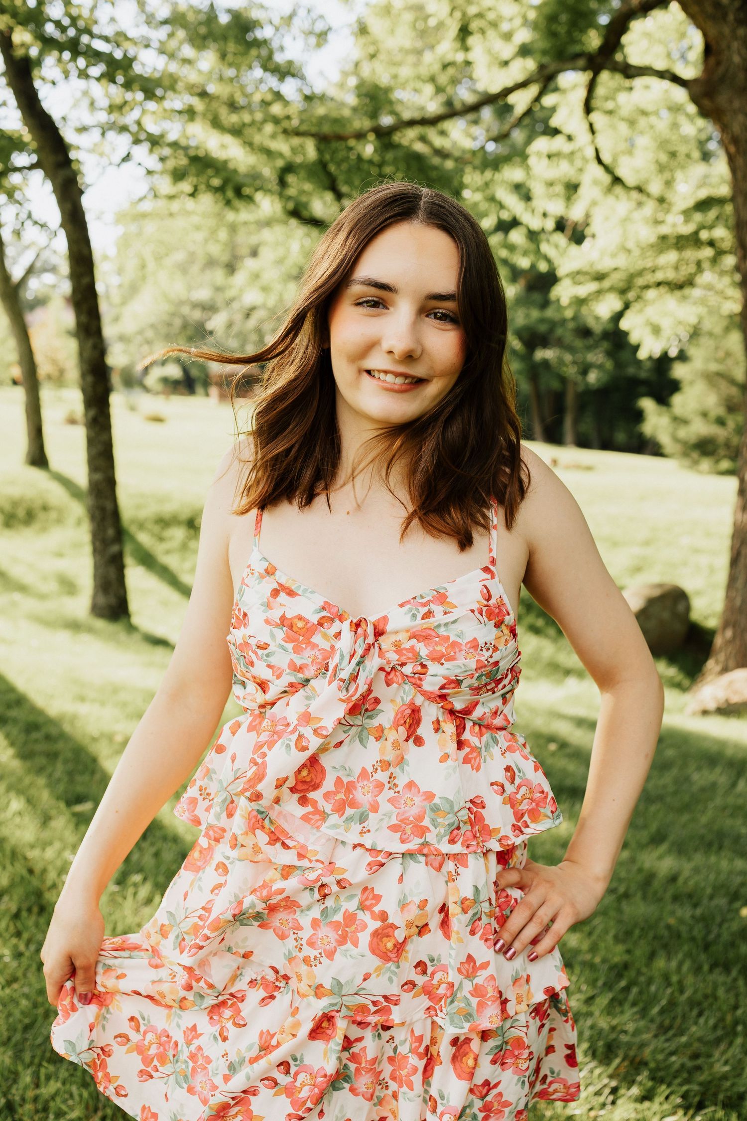 Young woman in floral sundress smiling outdoors in a lush green park on a sunny day.