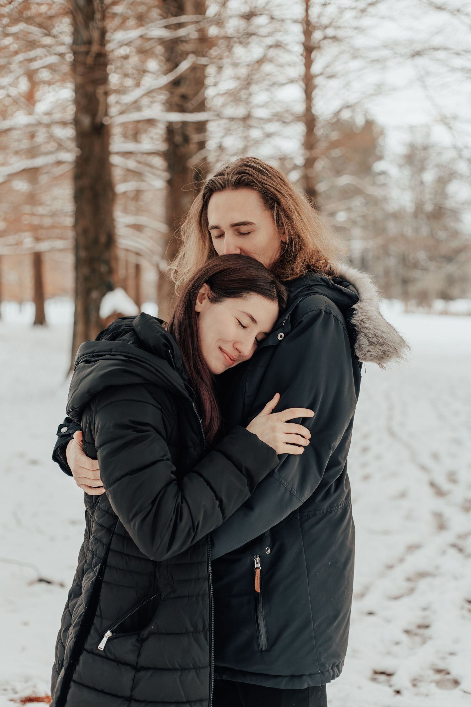 A couple embraces in winter coats against a snowy forest backdrop.